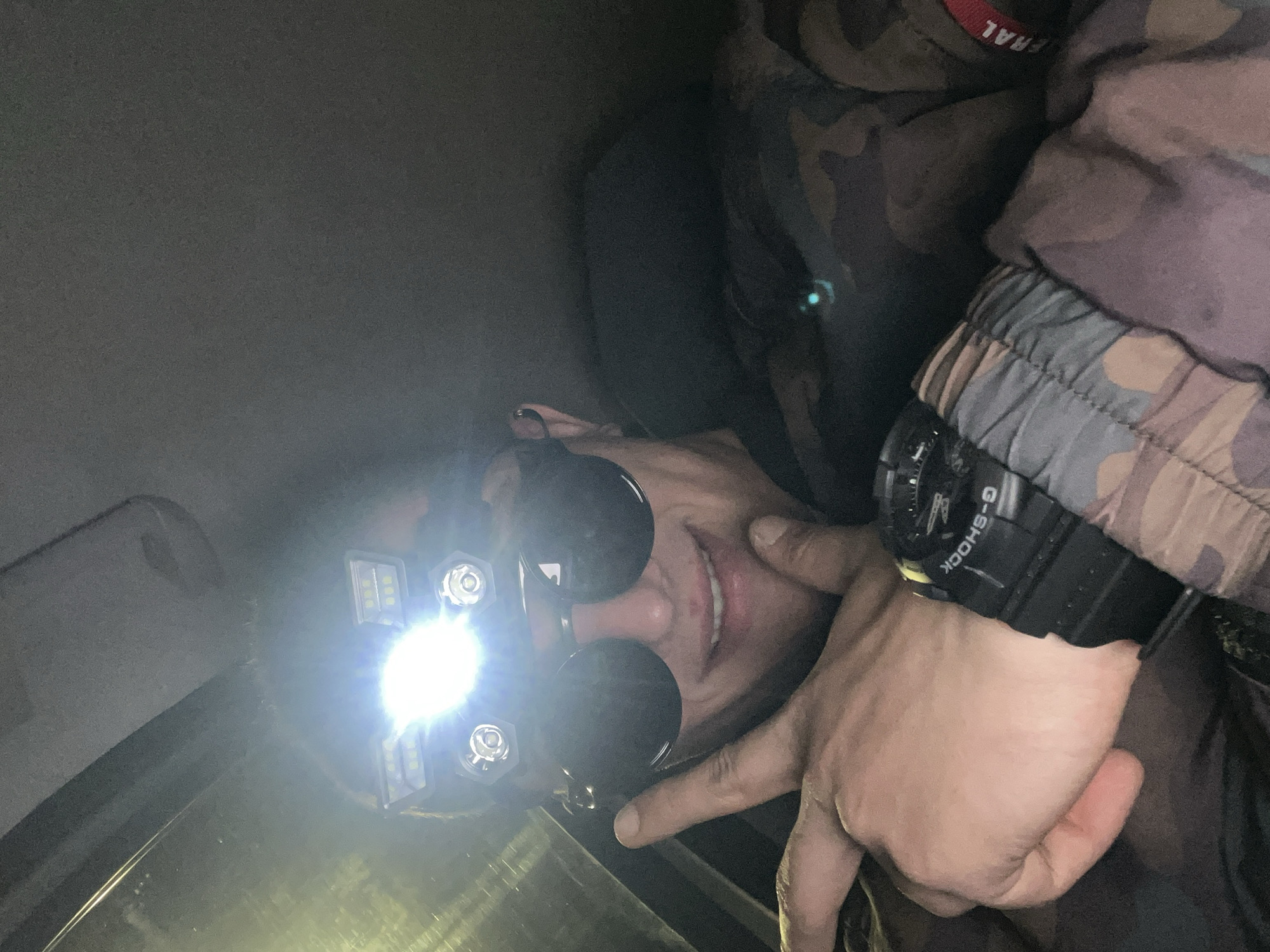 A person wearing a headlamp and sunglasses is illuminated by the bright light of the headlamp. The person has their hand near their face and is wearing a G-Shock watch. The individual is wearing a camouflage jacket. The location is indiscernible, but it is supposedly located in Plovdiv, Bulgaria. The image has been rotated.