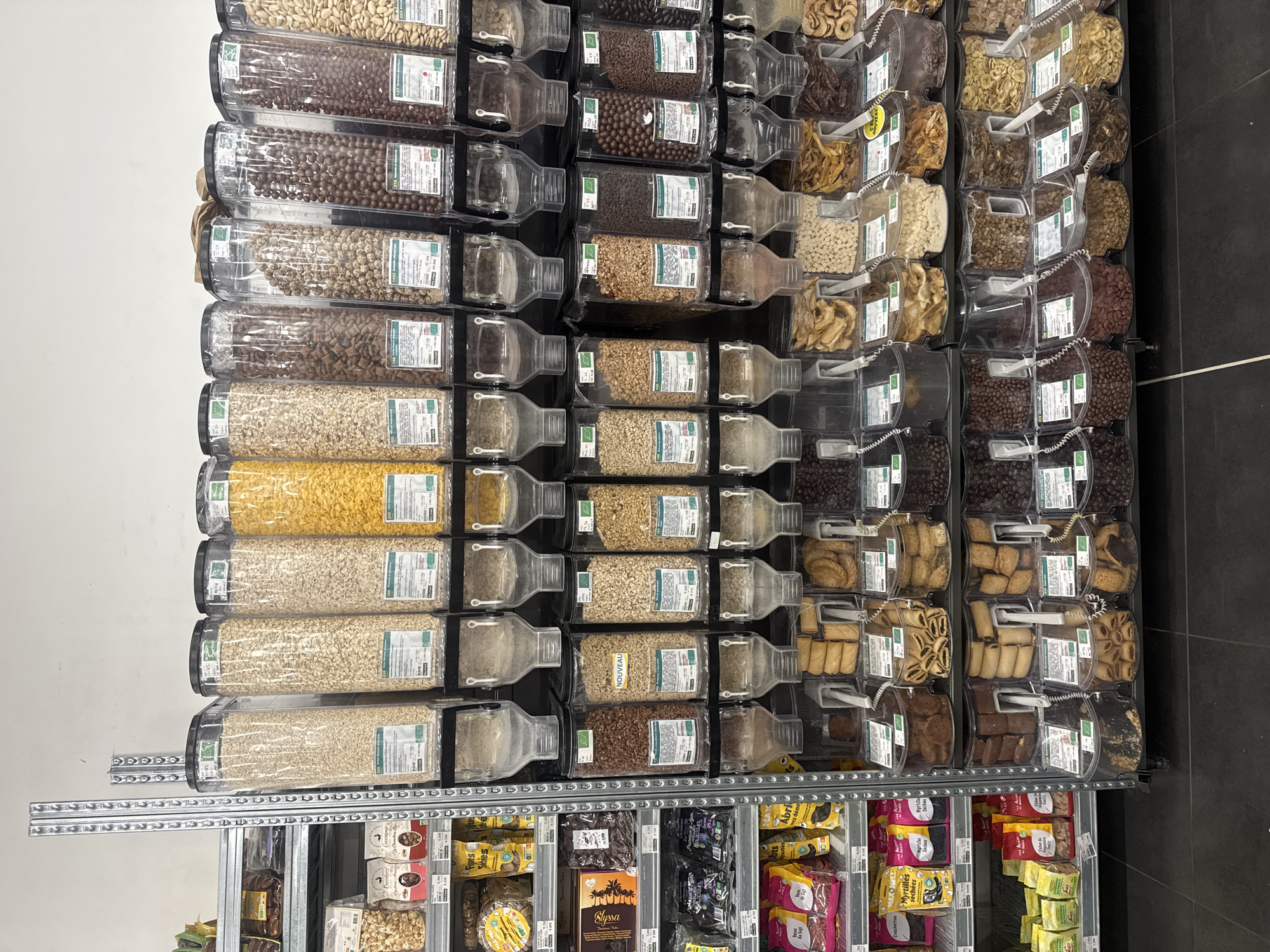This image shows a grocery store shelf in Paris, France. The shelves are stocked with various bulk food items, including grains, cereals, dried fruits, and nuts, stored in clear plastic dispensers. Below the dispensers, there are packaged food items such as dried fruits and chocolates. The environment appears to be a modern, well-lit retail space.