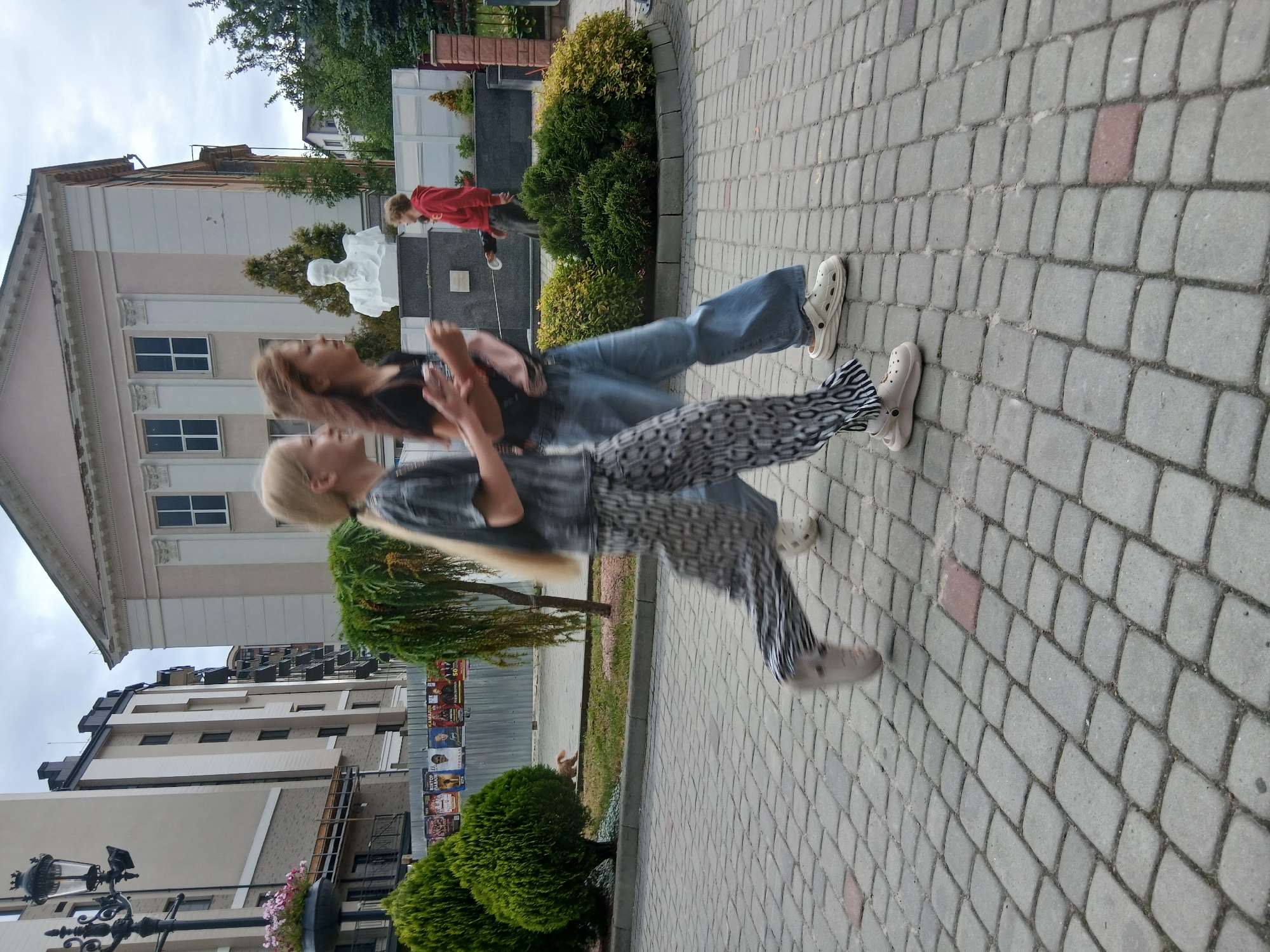 Two young females are walking on a cobblestone walkway in front of a building. The female on the left has blonde hair and is wearing a gray t-shirt with a black pattern and patterned pants. The female on the right has brown hair and is wearing a black t-shirt and blue jeans. Behind them, there is a large building with columns and windows, a bust of a man, and some trees. In the background, there are other buildings and a street. The sky is overcast. The location is the city of Truskavets, Ukraine.