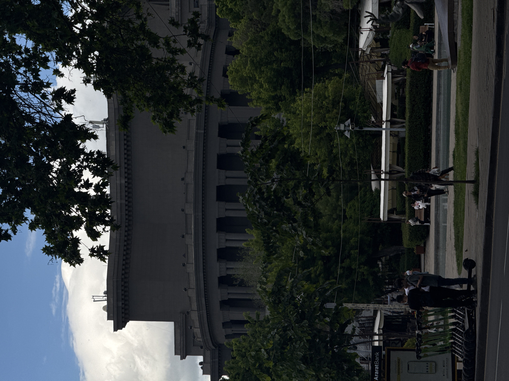 This photo shows a section of Yerevan, Armenia, with the Cascade monument in the background. The sky is partly cloudy. Trees obscure parts of the building. There are people visible on the sidewalk and near the fountain. An advertisement for Araratbank and a row of electric scooters can be seen in the foreground.