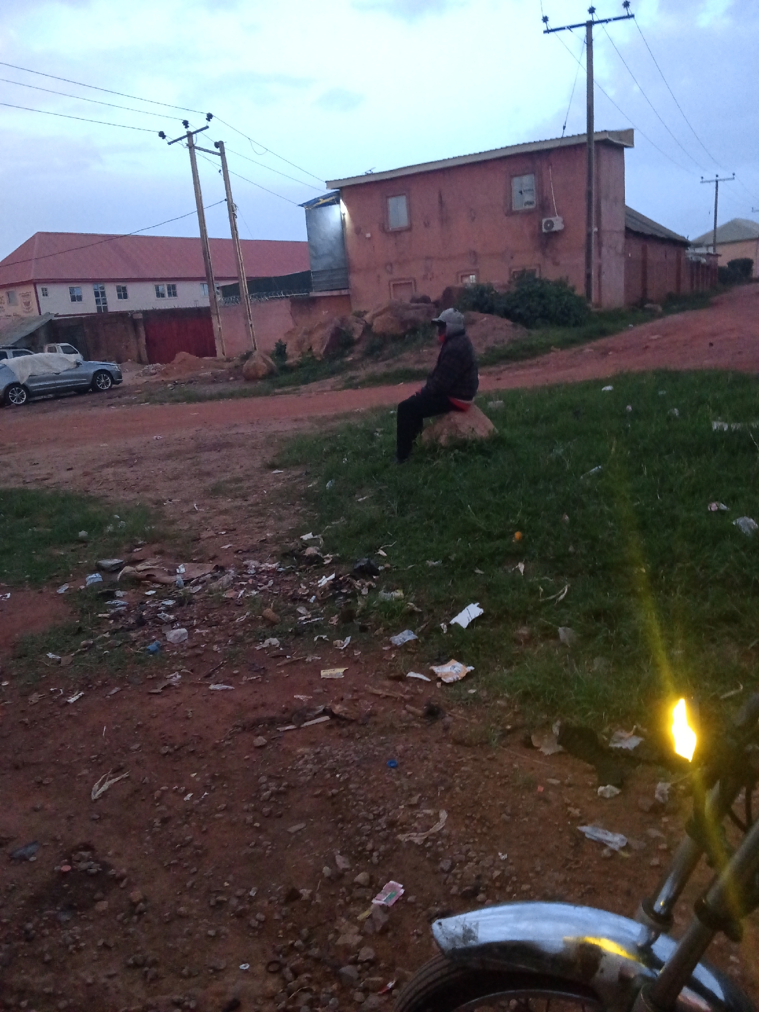 In Barakin Lerawi, Nigeria, a man wearing a dark jacket and a grey baseball cap sits on a rock on a grassy patch of ground. The ground is littered with various pieces of trash, including plastic bags, paper wrappers, and other debris. Behind the man, a red, two-story building with visible windows and an air conditioning unit is situated next to a power pole with electrical wires running across it. Another power pole is visible in the background. A large, red-roofed building with multiple windows is seen further in the background, to the left of the red building. Two vehicles are parked in front of this building, one of which appears to be covered with a grey tarp. The area is characterized by dirt roads and sparse vegetation. The lighting suggests it is either early morning or late evening. In the foreground, the front of a motorcycle is visible, with its handlebars and fender reflecting light.