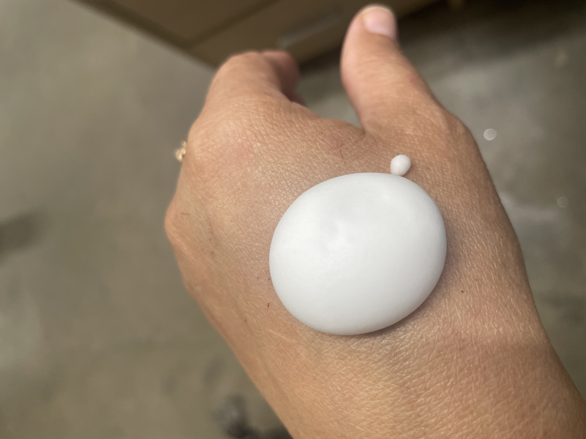 A close-up, overhead shot shows a person's left hand with a large dollop of white cream on the back of it. A tiny bead of the same cream is visible to the right of the larger dollop. The hand is resting on a textured, neutral-colored surface. A hint of a ring is visible on one of the fingers, as is a portion of a cardboard box in the background. The image was taken in the city of Tervolovo, Russia.
