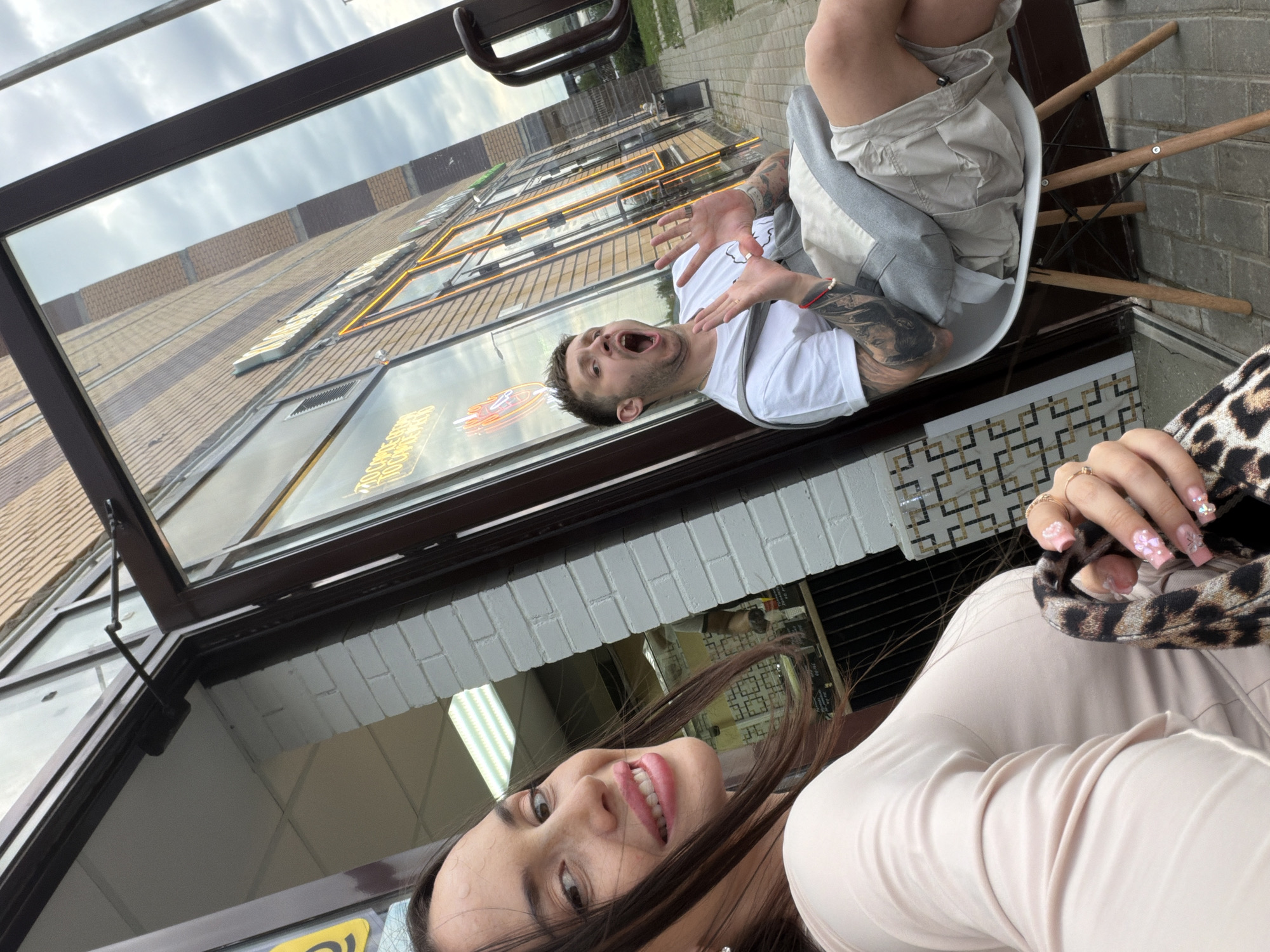 The image depicts an outdoor scene in front of a commercial establishment in Saint Petersburg, Russia, specifically identified as "КРАФТОВЫЙ ПАР" (Craft Vapor) at Prospekt Prosveshcheniya, 80 корпус 1.
In the foreground, a woman with long, dark hair is positioned slightly to the left, capturing a selfie. She is smiling, looking directly into the camera, and wearing a light-colored, long-sleeved top. Her right ear features a small, metallic stud earring. Her left hand, adorned with elaborate pink nails featuring 3D floral and gem designs, is holding a leopard-print object, possibly a handbag or scarf.
Reflected in the large glass storefront behind the woman, a man is visible seated in a white, modern chair with wooden legs, positioned outdoors on the paved sidewalk. He is wearing a white short-sleeved t-shirt and light-colored shorts. Both of his arms are heavily tattooed with dark designs. A grey shoulder bag or pouch is draped over his left shoulder. He has a red beaded bracelet on his left wrist. The man's mouth is wide open in an exaggerated expression, and his hands are raised with palms facing forward, as if in surprise or emphasis.
The storefront itself features dark brown framed glass panels, including an entrance door, outlined with bright orange neon lights. Within the shop, additional neon signs display Cyrillic text, including "КРАФТОВЫЙ ПАР" (Craft Vapor), "СИГАРЫ" (Cigars), and "КОФЕ" (Coffee), indicating the nature of the establishment. A dark-colored door closer mechanism is affixed to the top of the door frame. Adjacent to the storefront, a section of white painted brick wall is visible. The ground is paved with grey interlocking pavers. In the reflection of the glass, an overcast sky is visible, along with a distant building featuring alternating horizontal stripes of light and dark brown bricks, and a dark utility pole.