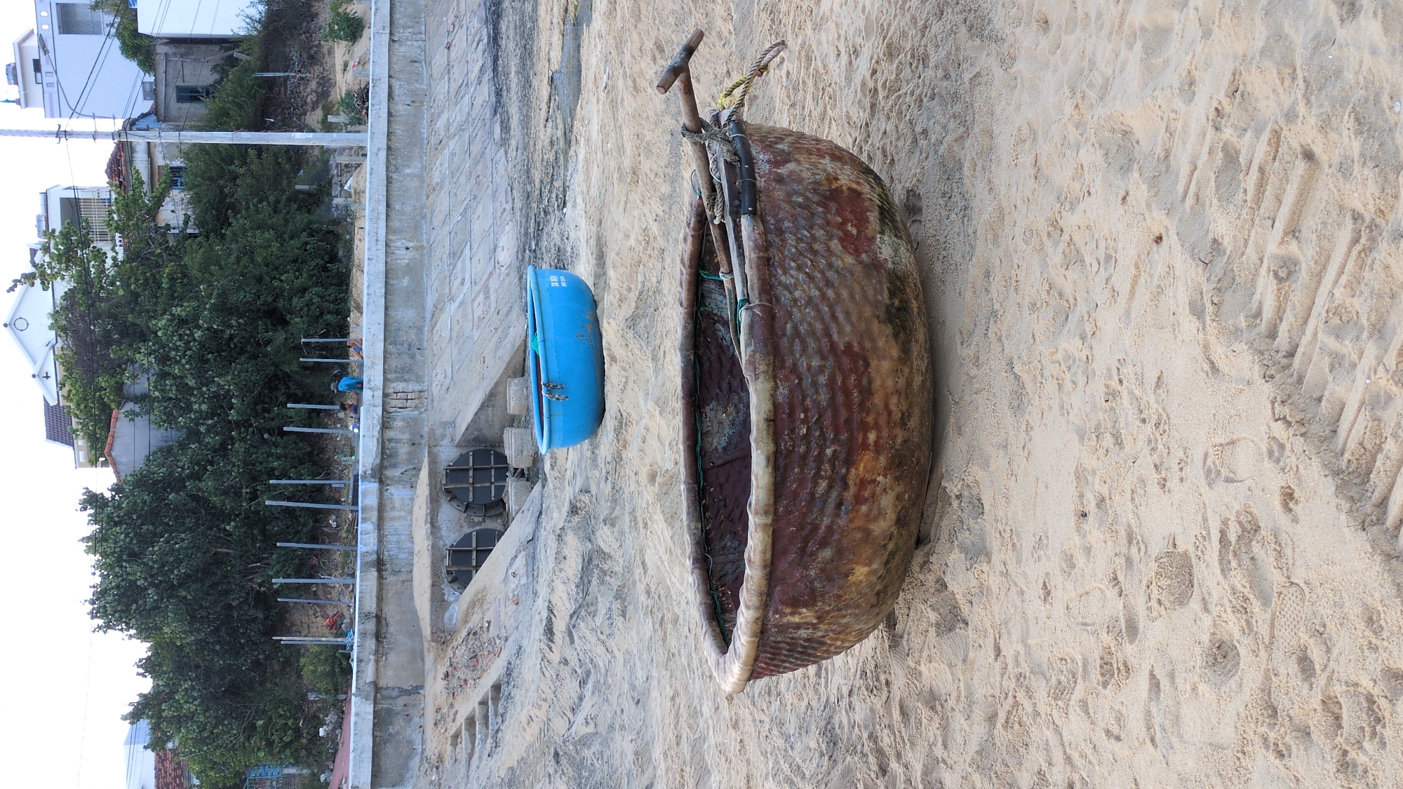 The scene captures a coastal area within East Quy Nhon Ward, Vietnam. In the immediate foreground, a large, weathered, reddish-brown coracle boat (thúng chai) made of woven material rests on light-colored sand, its interior containing two wooden poles secured with yellow and black ropes. The sand in this area displays numerous footprints and ripple patterns.
Midground elements include a smaller, bright blue plastic coracle boat positioned further back on the sand, featuring chains near its rim. Behind the boats, a substantial concrete seawall or embankment extends horizontally across the frame. This structure incorporates a sloped section with two large, circular drainage pipes equipped with grates, and partial concrete steps covered by sand are visible to the left of the pipes. Tire or drag marks are discernible in the sand leading towards the seawall.
The background consists of a dense treeline on a raised bank situated directly behind the embankment. Above the foliage, several residential buildings, primarily white with some red-tiled roofs, are visible against an overcast sky. One white building prominently features a cross on its roof. A utility pole with wires stands on the far right. On top of the concrete embankment, three individuals are present: one wears a light blue shirt, and two others are in darker attire, appearing to be resting or engaged in an unspecified activity near several grey vertical posts.