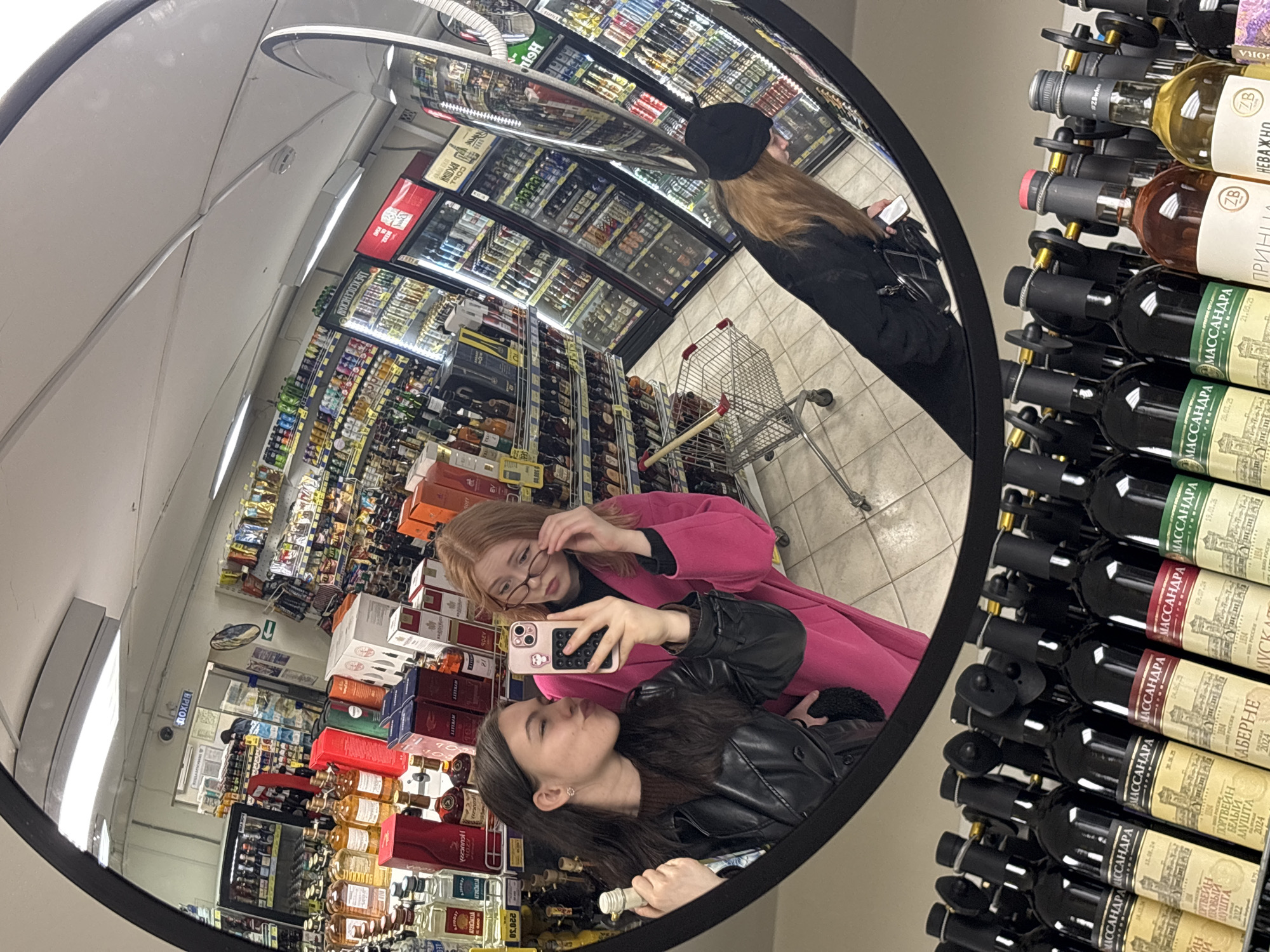 A wide-angle, circular reflection in a convex security mirror captures a scene inside a supermarket or liquor store in Perm, Russia. Two young women are the main subjects, seemingly taking a selfie. The woman in the foreground has dark hair and wears a black leather jacket; she holds a smartphone with a pink case, angled towards the mirror, and has a slight pout. Behind her, a woman with reddish-blonde hair and glasses, wearing a pink coat, adjusts her spectacles while looking towards the camera.
The mirror's reflection shows extensive shelves filled with bottles, predominantly wine and spirits. Refrigerated display cases holding various beverages are visible in the background. Further back in the reflection, another person with long brown hair and a black hat can be seen pushing an empty shopping cart. The store has bright, artificial lighting.
Visible Cyrillic text on bottle labels includes "МАССАНДРА" (Massandra), "ПОРТВЕЙН" (Portwein – Port wine), "БЕЛЫЙ АЛУШТА" (White Alushta), "КАБЕРНЕ" (Cabernet), and "МУСКАТ" (Muscat). Other labels show "НЕВАЖНО" (Nevazhno), "ПРИНЦИ" (Prince), "ZB", and "#ZBwin," along with various dates like "09.07.24" and "1854." A sign with "ПРИХОД" (Prikhod - Entrance/Arrival) is also faintly visible in the background. The overall scene depicts a casual moment of two friends in a retail setting, using an unconventional surface for a photo.