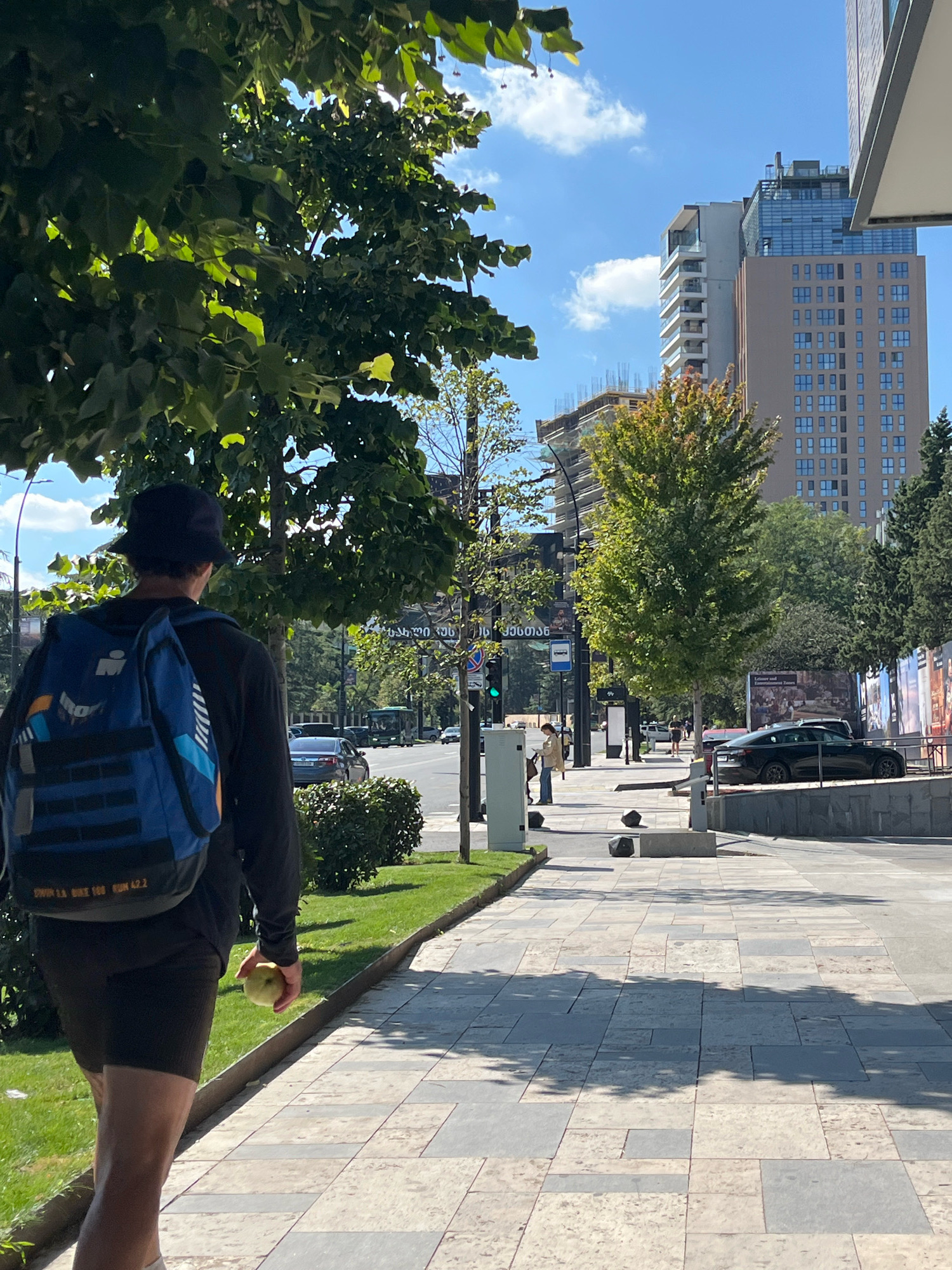 In Tbilisi, Georgia, a person wearing a dark bucket hat, a dark long-sleeved shirt, and dark shorts walks on a sidewalk. They are carrying a blue backpack with orange and white accents, and holding a light-colored apple in their right hand. The sidewalk is paved with large, rectangular tiles in varying shades of beige and gray, and is bordered by a grassy area on the left. Beyond the grass, trees with green leaves line the street.
In the background, several modern buildings are visible. One tall building is light brown with many windows, and another is taller with blue-tinted windows and a modern design. Traffic is present on the street, with cars and a bus visible. The sky is blue with scattered white clouds. The scene appears to be a sunny day in an urban environment.