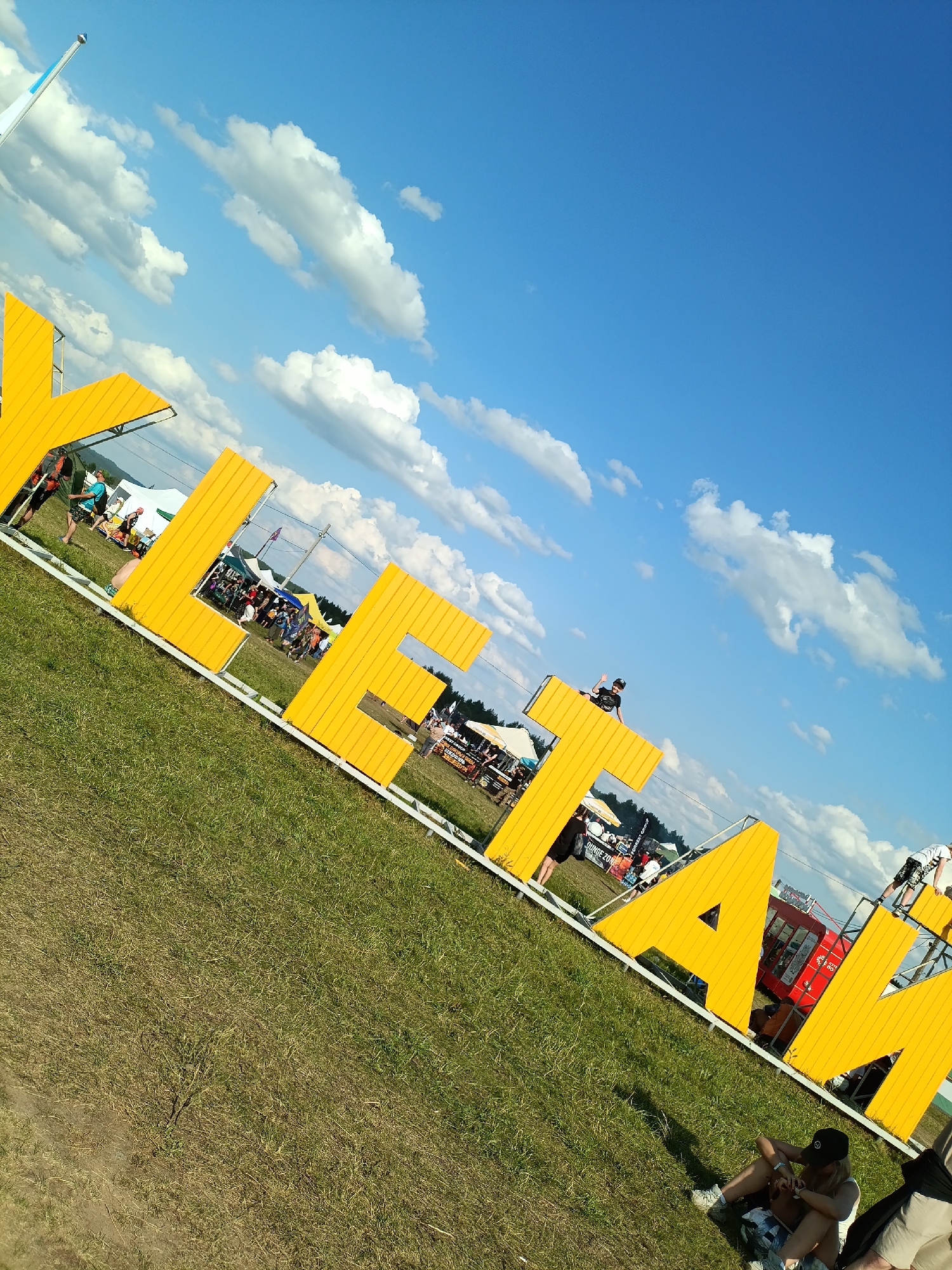 The image shows large, yellow, three-dimensional letters spelling out "YEKETAM" in Russian, likely a festival or event name. The letters are positioned outdoors on a grassy field under a bright blue sky with scattered white clouds. Several people are visible in the background, some walking, others standing near tents or small structures. One person is sitting on top of the letter "T", waving. In the foreground, to the right, a young woman is sitting on the grass, looking down at her phone, wearing a black cap. To the right of her, another person's legs are visible. The overall scene suggests an outdoor event or festival atmosphere. The location is identified as Lagunovo, Russia.