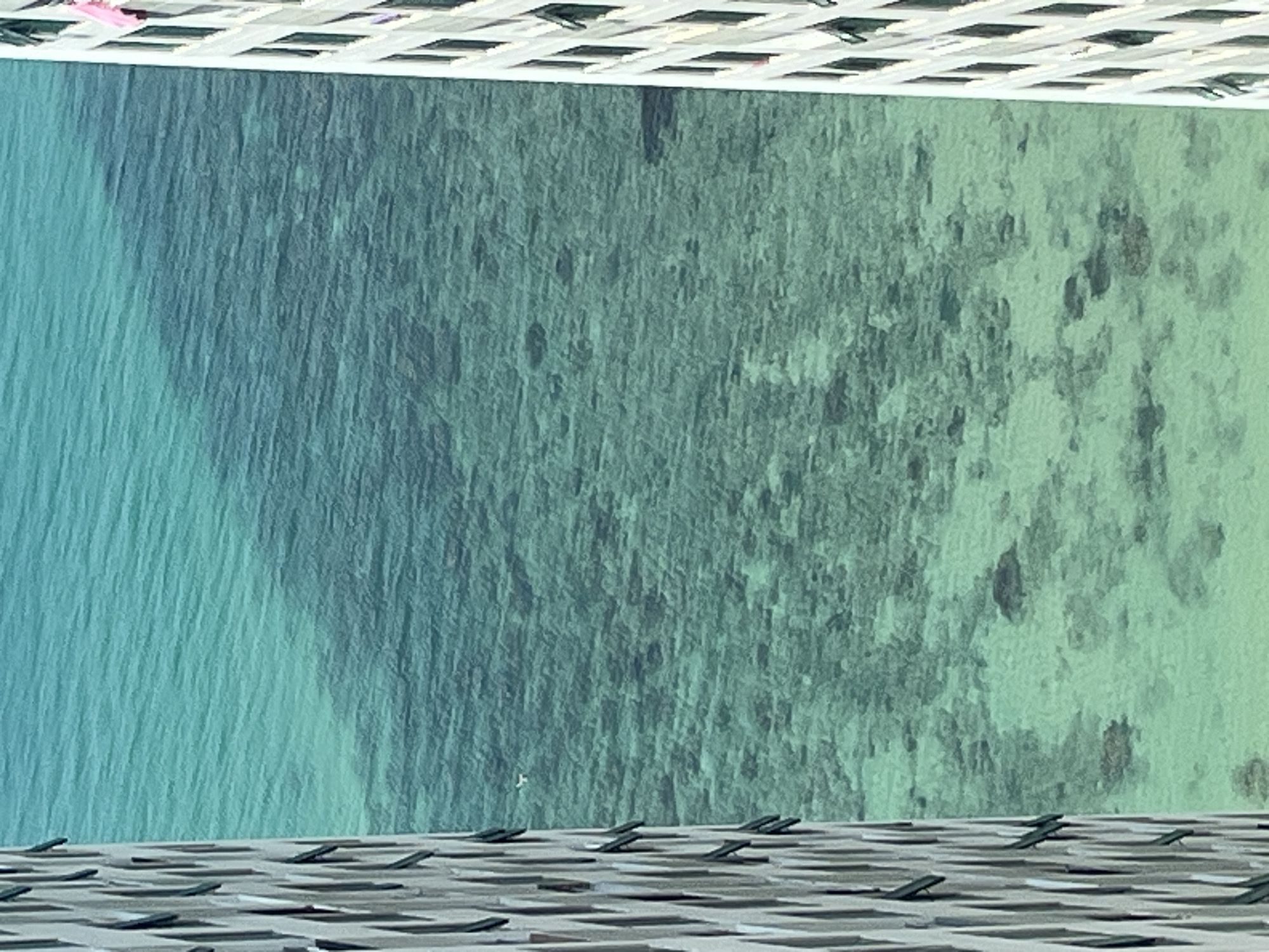 The image depicts an aerial view of a body of water, likely the ocean, with varying shades of blue and green, indicating different depths or features beneath the surface. Above and below the water, a grid-like pattern suggests a structure, possibly a roof or a platform, from which the image was taken. There is no indication of people, subjects, objects, or interactions. The location is given as Bac Nha Trang Ward, Vietnam.