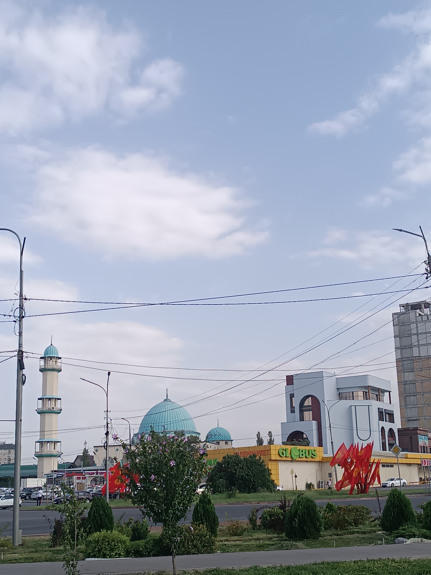 The image captures a street view in Bishkek, Kyrgyzstan, featuring a prominent mosque with a tall minaret and a large turquoise dome. To the right of the mosque, a modern white building with distinctive arched windows is visible, bearing the "GLOBUS" supermarket logo. A cluster of red flags with a yellow emblem, likely representing Kyrgyzstan, is displayed in front of the supermarket. The scene is framed by utility poles and overhead wires, common in urban environments. The sky above is partly cloudy, with a few birds visible in the distance. Greenery, including trees and bushes, lines the foreground, suggesting a well-maintained public space. Several cars are parked or driving on the street, indicating a typical day in the city. The overall impression is a blend of traditional Islamic architecture and contemporary commercial development.