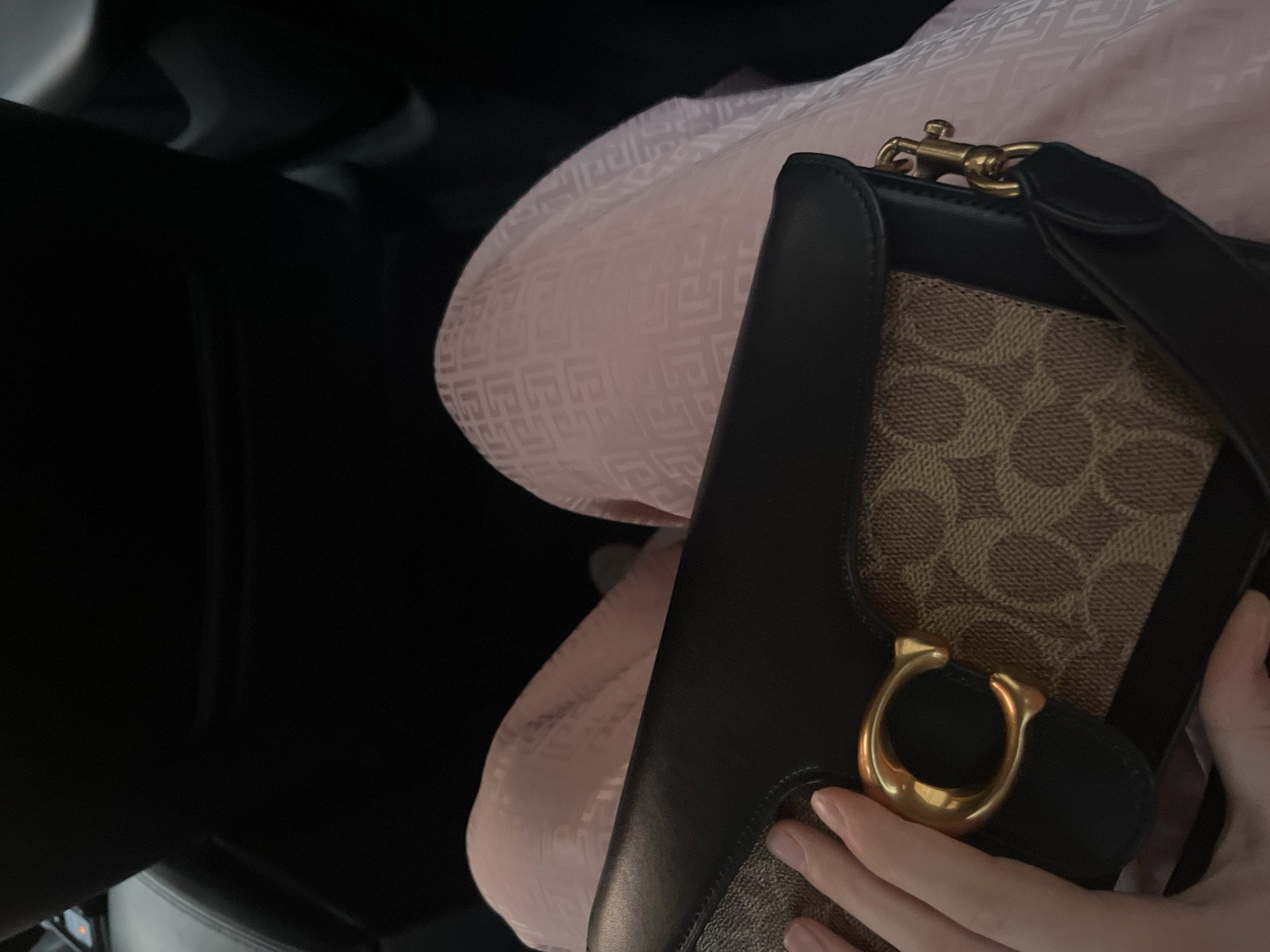 The image depicts a Coach Tabby shoulder bag with a Signature canvas and leather trim. The bag is positioned on what appears to be a patterned, light pink garment. A hand is visible, partially obscuring the bottom of the bag. The background shows a black car interior. The location is unknown and there is no indication of it being Moscow, Russia.