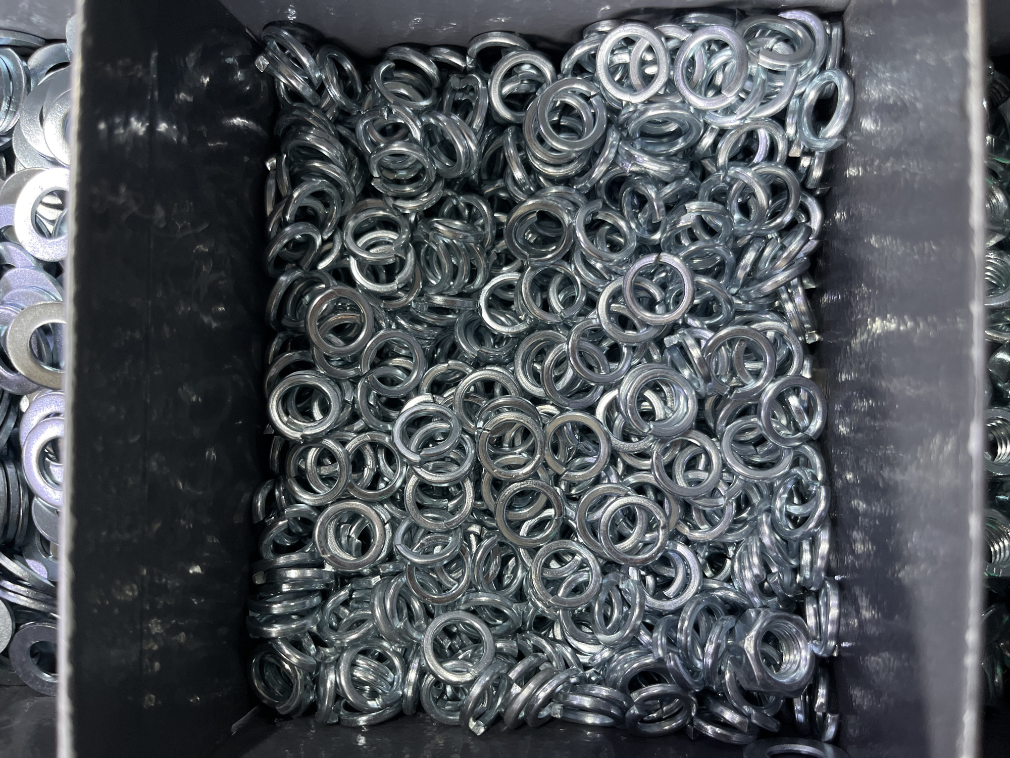 This media file shows a close-up, overhead view of a dark gray bin filled with many metal washers. The washers are predominantly shiny silver and appear to be of the split-ring or lock washer variety, characterized by their helical shape and gap. They are piled high within the bin, creating a textured surface of overlapping circles.
The lighting suggests an indoor environment, possibly a workshop or storage area, with the light source coming from above and slightly to the left, casting subtle shadows among the washers. The bin itself appears to be made of plastic or a similar material, with some scuff marks and reflections visible on its interior walls.
The image is focused on the washers, and there are no people or other significant objects visible. The activity taking place is simply the static arrangement of these hardware components within their container. There are no discernible time of day, weather, or strong emotional cues. The scene is purely an observation of manufactured items. No visible text is present in the image.