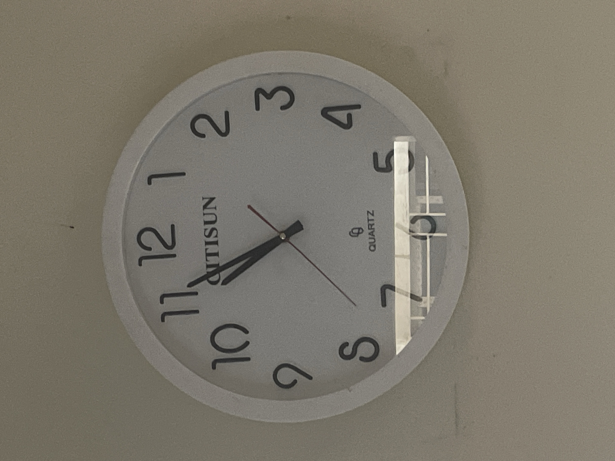 A white-cased analog clock is mounted on a light gray wall. The clock face has black numerals marking the hours and the brand name "ANTISUN" above the center. The hour hand is pointing just past the number 7, while the minute hand is closer to the 12. A red second hand is visible. A reflection is visible on the clock face. The image does not provide information regarding the location of the subject in Dhaka, Bangladesh.