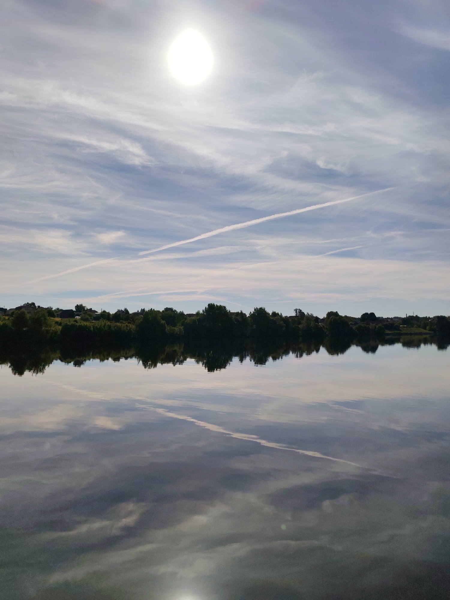The image depicts a static landscape scene in the city of Unknown, Unknown, primarily composed of a sky, a treeline with distant structures, and a body of water. The upper half features a light blue sky with diffuse, wispy white clouds and several distinct white contrails, one of which traverses diagonally from the lower-left to the upper-right of the sky section. A bright, overexposed white orb, identified as the sun, is prominent in the upper-right quadrant. The horizon line is defined by a dark green treeline, behind which multiple dark-roofed structures, indicative of buildings, are partially visible. The lower half of the image consists of a calm body of water that functions as a reflective surface. This water mirrors the sky, clouds, and contrails, reproducing the diagonal contrail and the diffuse cloud patterns with muted tones. A bright, less defined reflection of the sun is observable in the lower-right quadrant of the water. No individuals, animals, or explicit actions are depicted.