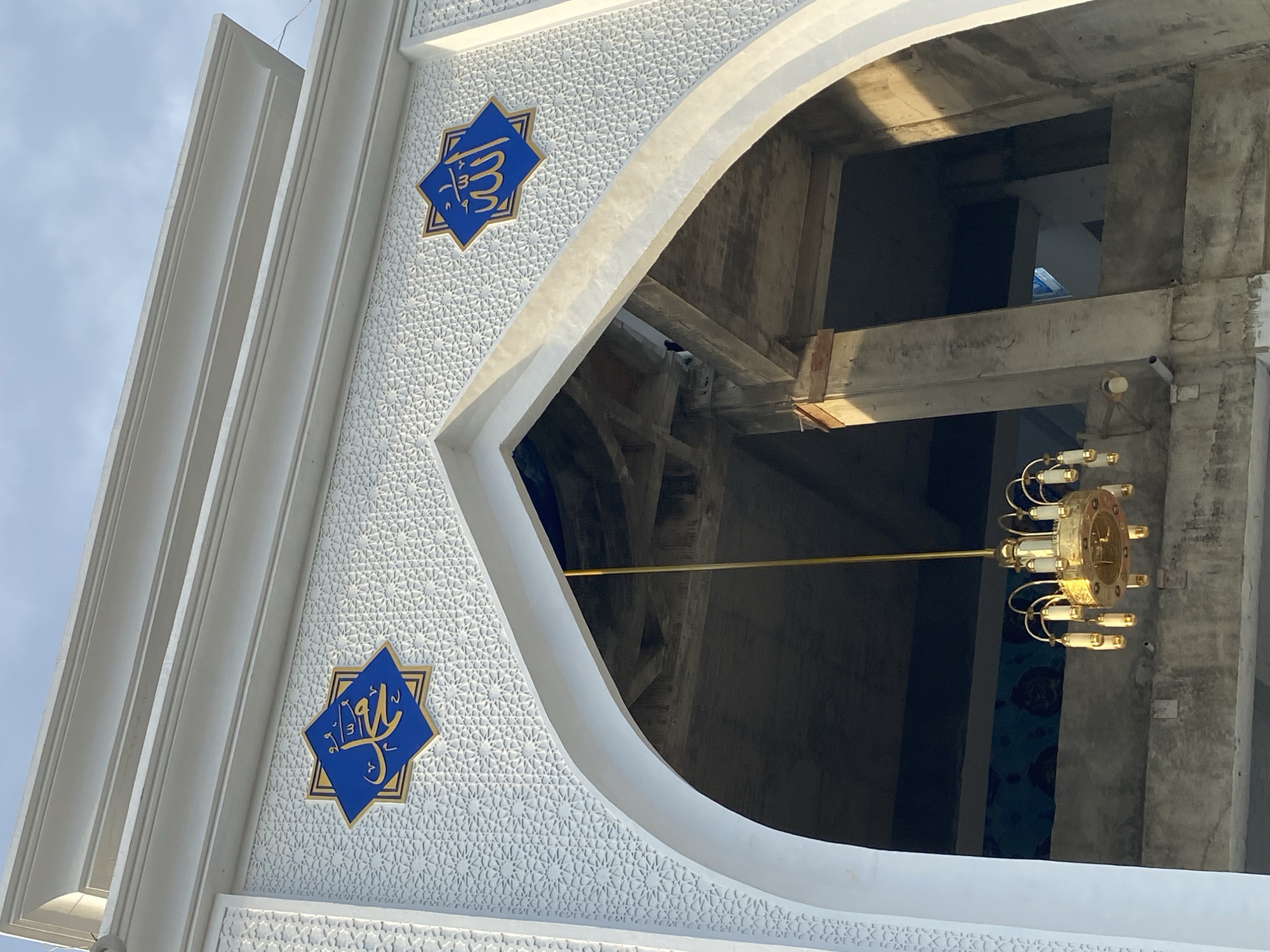 The image shows an upward view of the exterior of a mosque under construction in Lhokseumawe, Indonesia. The mosque features white decorative architectural elements, including a prominent archway. Two blue star-shaped plaques with gold Arabic calligraphy are mounted on the white facade above the arch. One plaque contains the name "Muhammad" and the other contains the word "Allah". A gold chandelier hangs from the ceiling within the archway, and the interior of the mosque appears to be unfinished, with exposed concrete pillars and ceiling structures. The sky is visible in the background.