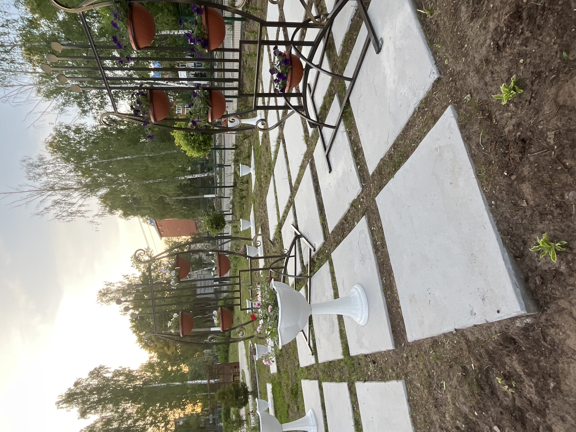 The image depicts an outdoor scene in what appears to be a garden or landscaped area, possibly in Chaikovsky, Russia, given the prompt. The scene is viewed from an elevated position, looking down at an angle. A series of square concrete paving stones are laid in a grid-like pattern, with patches of soil and grass visible between the stones. Along this pathway, there are decorative metal structures, likely plant stands, featuring hanging flower pots. Some of the pots contain purple flowers. Interspersed among these are white, pedestal-style planters, also holding flowers. The background features trees, some of which appear to be birch trees, and glimpses of buildings, including what may be a brick structure. The sky is visible in the upper left, suggesting it is either early morning or late afternoon.