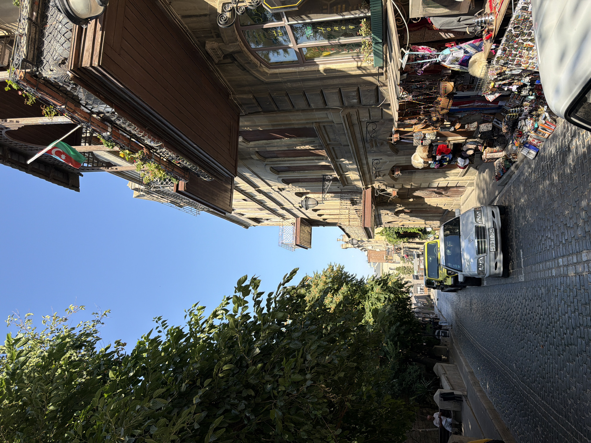In Sabail Raion, Azerbaijan, a cobblestone street is lined with buildings and trees under a clear blue sky. The street is narrow and slopes slightly downhill, with cars parked along the right side. On the left, a person sits on a bench beneath a large tree. The buildings on the right are several stories high, with ornate balconies and windows. One building features a shop displaying various handicrafts, including bags, jewelry, and souvenirs. An Azerbaijani flag flies from a balcony on an upper floor. The scene is illuminated by bright sunlight, casting shadows on the street and buildings.