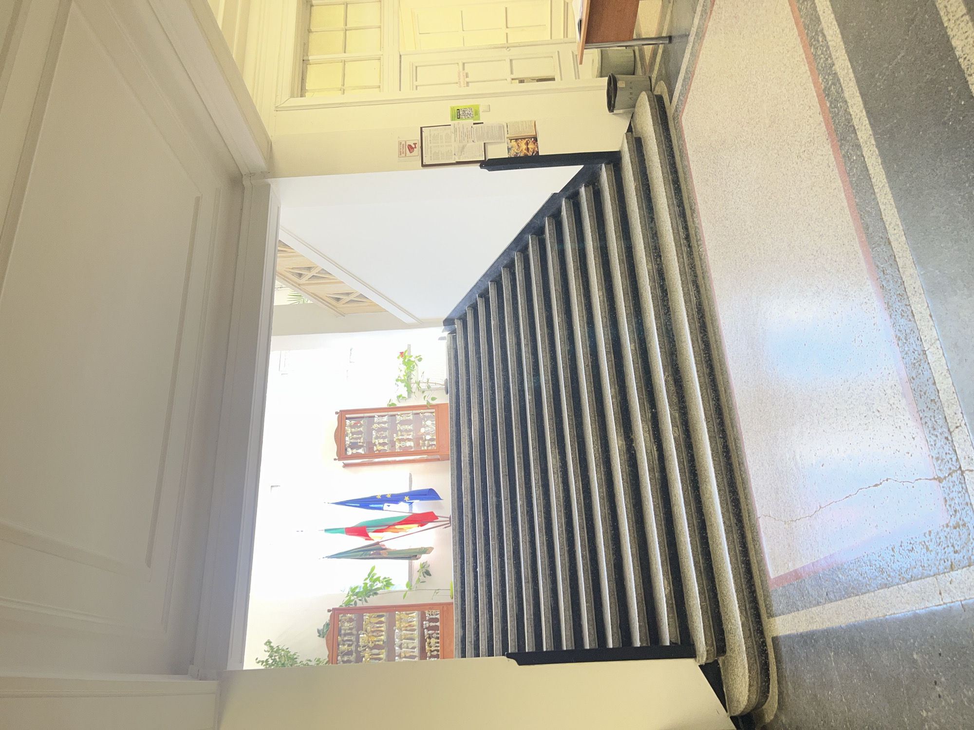 The image shows a hallway with white walls and a gray and white patterned floor. A doorway on the left leads to a bright room with two display cases and three flags hanging on the wall. The flags are blue, red, and green. A black railing is on the right side of the hallway. There are windows at the far end of the hallway. A bulletin board with papers is above the railing. The location is likely in Sofia, Bulgaria, given the flags.