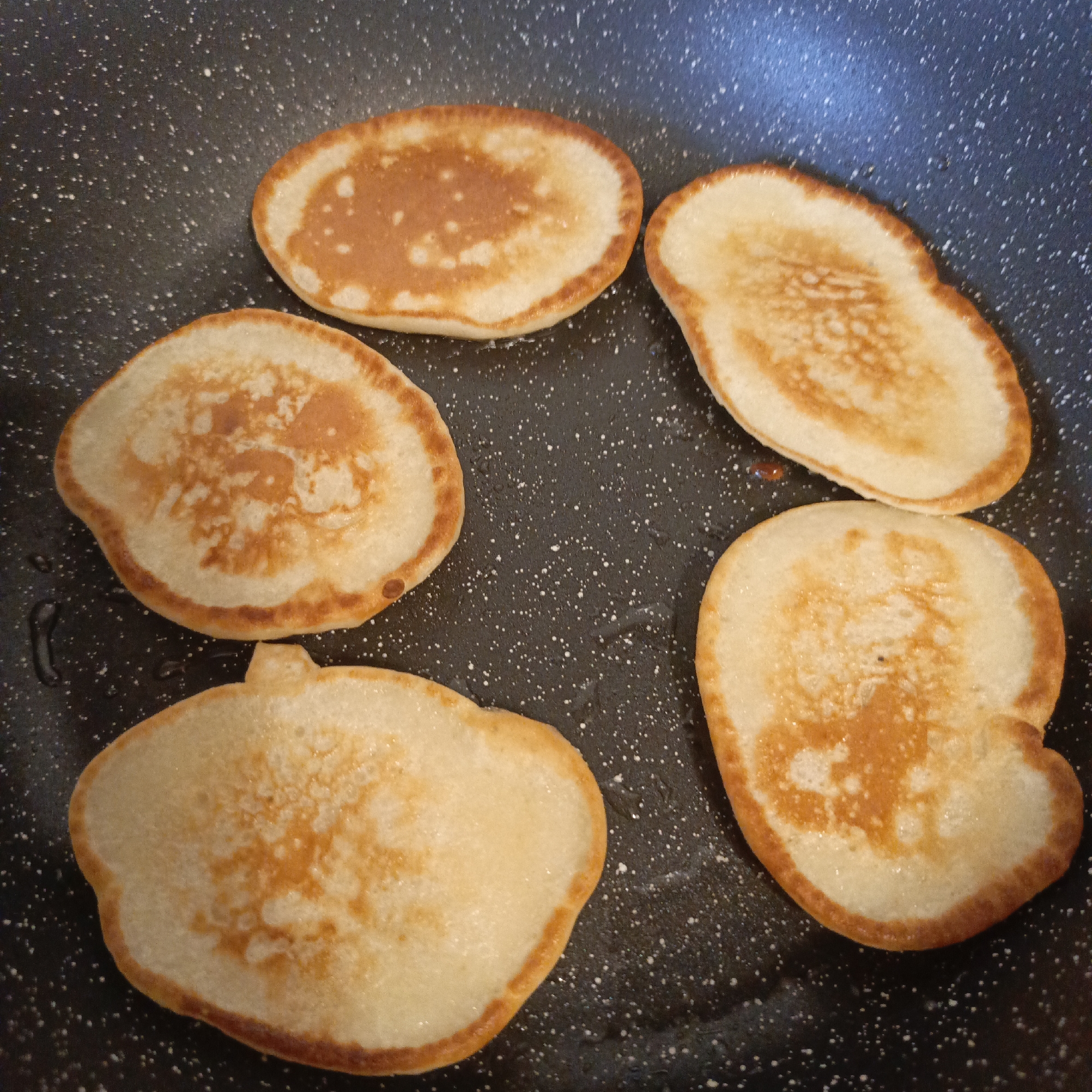 Five golden-brown pancakes are cooking in a dark, speckled non-stick frying pan, viewed from directly above. The pancakes are arranged in a circular pattern within the pan, with one pancake at the top, one to the top right, one to the bottom right, one at the bottom, and one to the mid-left. They vary slightly in shape and size, but all have characteristic golden-brown edges and lighter, fluffy centers with some areas of browning. Some small bubbles are visible on the surface of the pancakes, indicating they are actively cooking or have recently been cooked. The pan itself is dark gray or black with numerous small white speckles, suggesting a non-stick coating. There are faint reflections of light visible on the pan's surface, particularly in the upper right and top left areas. No people or text are visible in the image. The scene suggests an indoor kitchen environment, likely on a stovetop, capturing a moment of breakfast preparation.