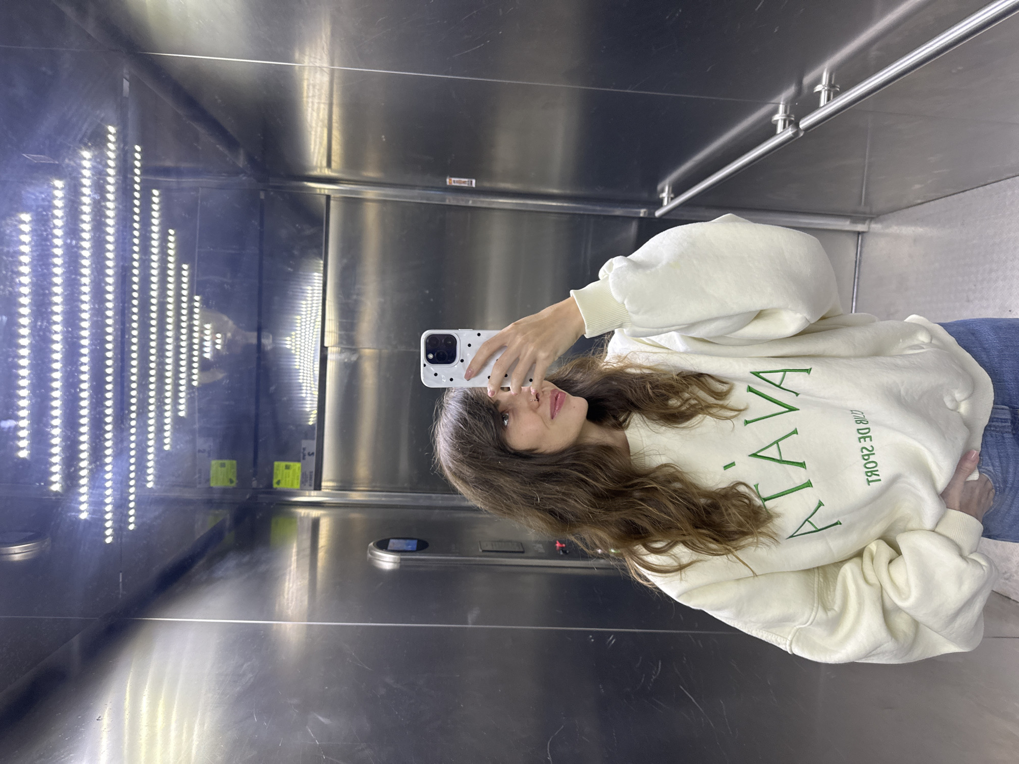 A young woman with long, wavy brown hair is taking a selfie in an elevator. She is wearing a cream-colored sweatshirt with green lettering that says "ATAVA" and "CLUB DE SPORT" in reverse. Her right hand is holding her phone, which has a white case with black polka dots, and her left hand is resting on her hip. The elevator has stainless steel walls and ceiling, with a row of bright LED lights on the ceiling. The floor appears to be metallic as well. The elevator is located in Tnuvot, Israel.