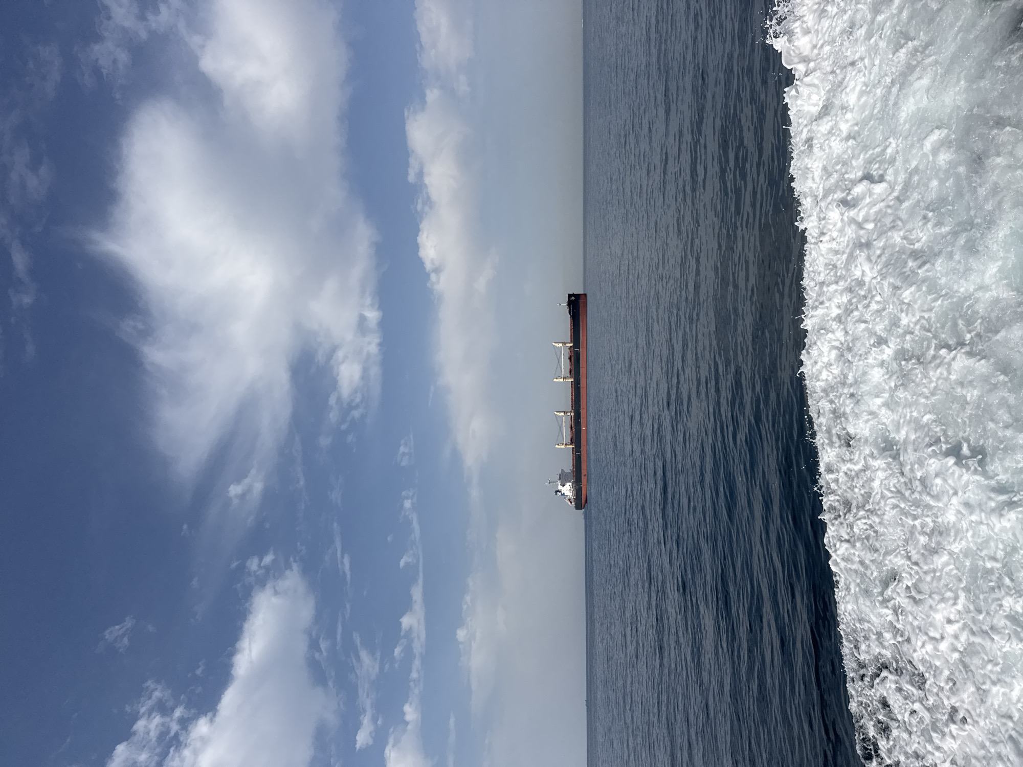 A cargo ship with a red hull and a dark deck is sailing on a vast, calm blue sea under a bright, partly cloudy sky. The ship is positioned in the center of the frame, at a distance from the viewer. The foreground of the image shows the white wake of a moving boat, indicating the perspective is from a vessel. The horizon line is clearly defined, separating the sea from the sky. The sky is a vibrant blue, with scattered white cumulus clouds. There is a faint haze in the distance along the horizon. The overall scene depicts a nautical environment.