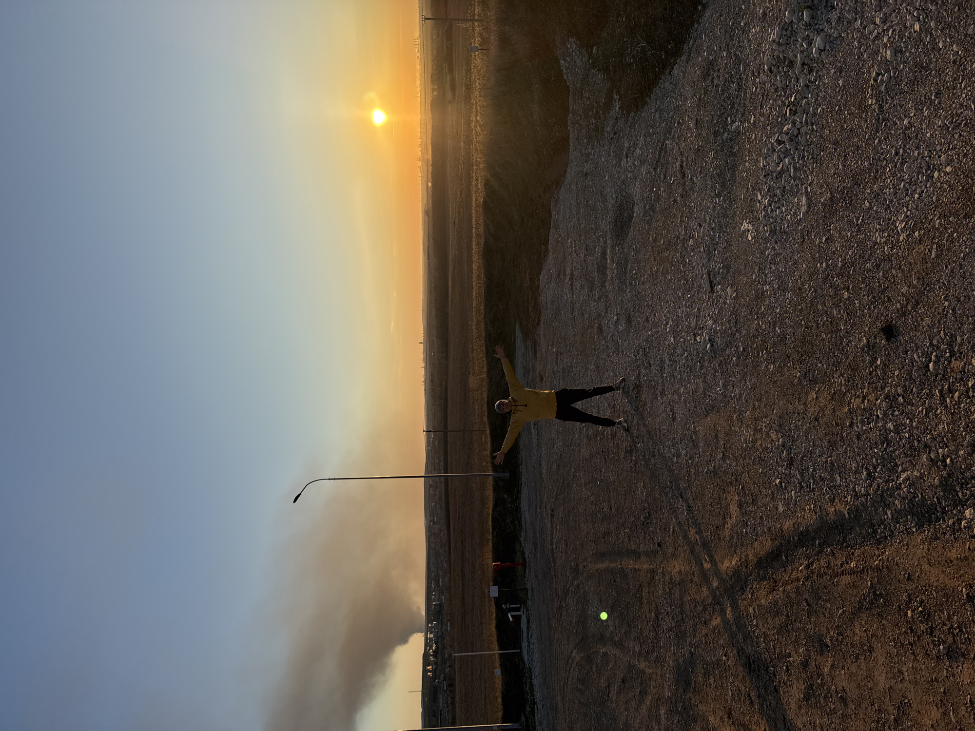 A person with their arms outstretched lies on a gravel-covered ground, with a field stretching towards the horizon. The sun is setting in the distance. A street light stands to the left. A cloud of smoke is visible in the distance.
Based on the request for the location in Kochkino, Russia, and the presence of smoke, the scene could possibly depict a field or rural area in Kochkino, Russia.