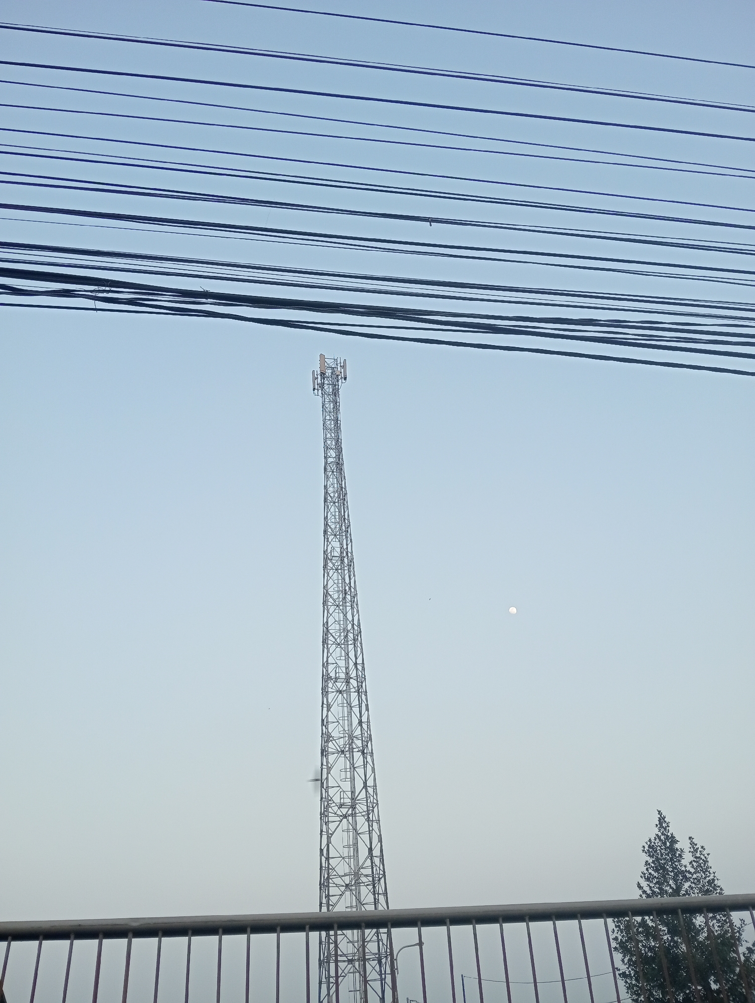 A transmission tower is shown against a clear sky, with numerous power lines running horizontally across the top of the frame. A faint, round object, possibly the moon or the sun obscured by haze, is visible in the sky to the right of the tower. In the foreground, a metal railing with vertical bars runs along the bottom of the image. A few scattered trees are visible to the far right, and a small bird is in flight near the transmission tower. The scene is an outdoor environment, likely in Waluyojati, Indonesia, characterized by its infrastructure and open sky.