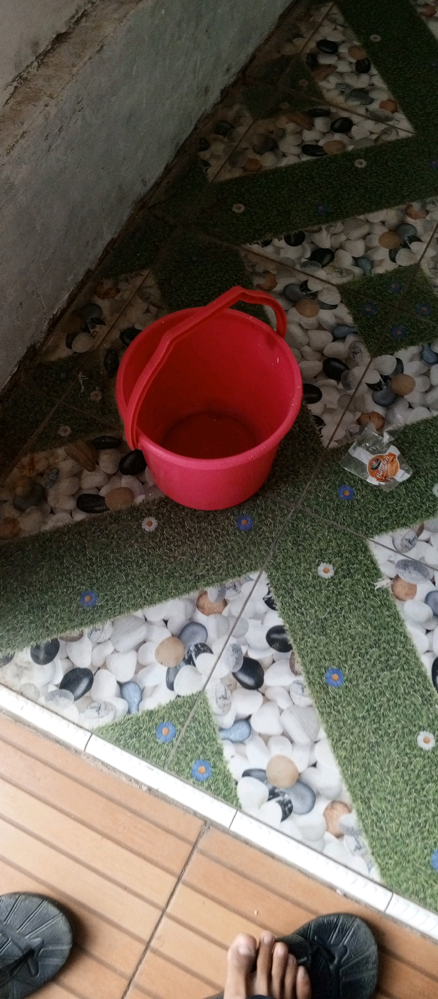 A red plastic bucket with a handle sits on a decorative tiled floor. The floor design features green grassy areas interspersed with white pebbles and small blue and white flowers. To the left, a concrete wall is visible, and in the foreground, two feet wearing black flip-flops are partially in the frame. A small piece of candy wrapper is also on the floor near the bucket. The scene appears to be located outdoors, possibly in a yard or patio area, in Karawang, Indonesia.