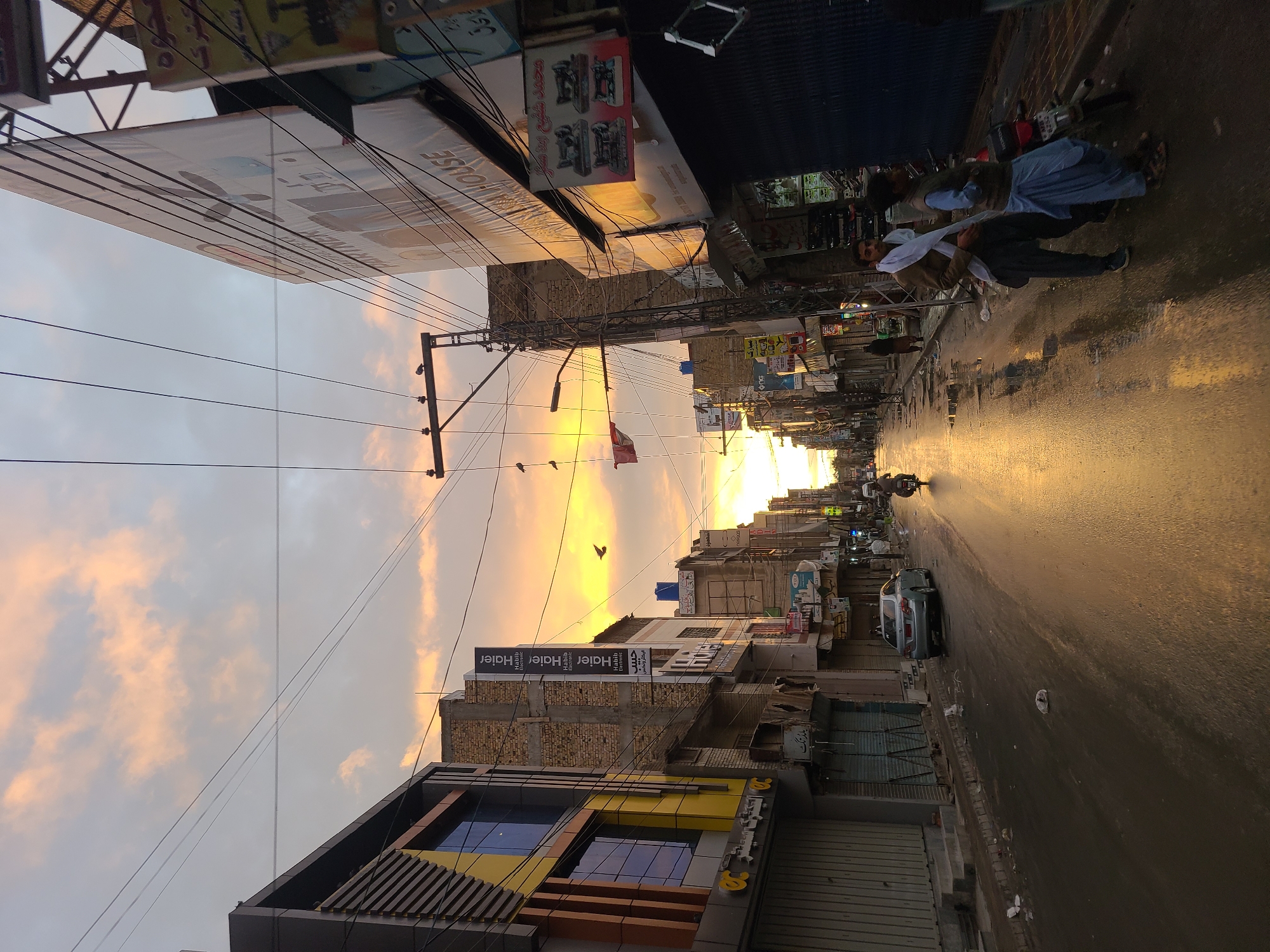 The image depicts a street scene in Quetta City Tehsil, Pakistan, at dusk. The road is wet, reflecting the golden light of the setting sun. Buildings line either side of the street, with a mix of brick and more modern facades. Signs and advertisements are visible on the buildings, including a Haier sign. Power lines crisscross overhead. There are people on the right sidewalk. Vehicles, including a car and a motorcycle, are on the road. A red flag hangs between the buildings. Birds are flying in the sky.