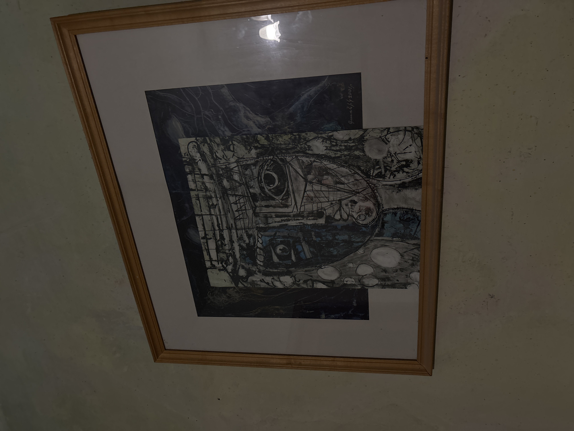 The image shows a framed artwork hanging on a wall. The artwork consists of two overlapping pieces. The background piece is a dark blue and black abstract painting with some visible brushstrokes. The foreground piece is a black and white abstract drawing with an apparent eye-like shape formed by lines and circles. The drawing also includes grid-like structures. The artwork is framed in a simple wooden frame. The wall is pale and shows some textures. Due to the image content, it is not possible to identify the location in Yerevan, Armenia.