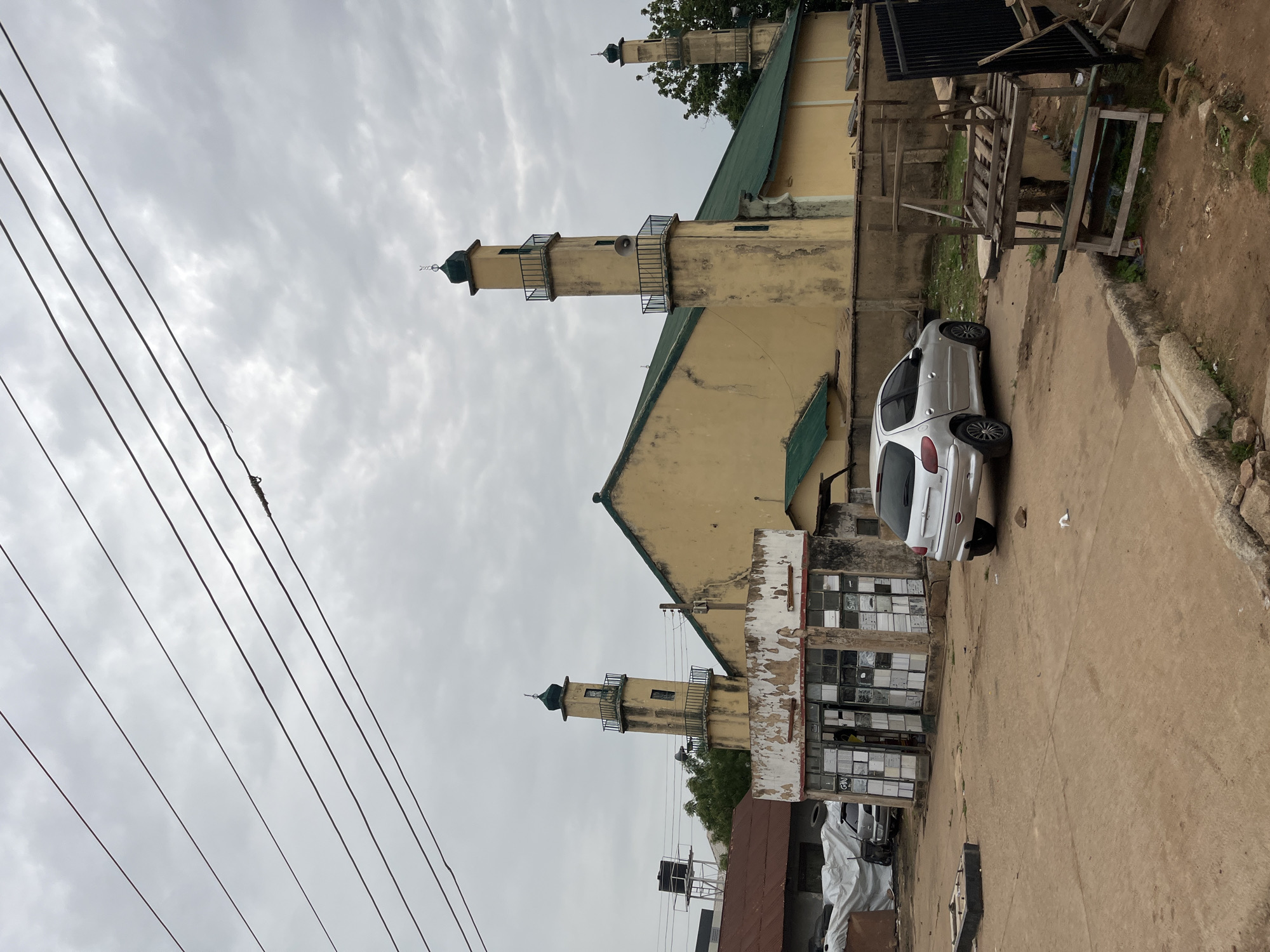 This is a medium shot, outdoor, full-frame photograph of a mosque with multiple minarets, located in Bukuru, Nigeria. The mosque is painted yellow with green roofs and features at least three prominent minarets, each topped with a green dome. In the foreground, there is a silver Peugeot car parked on a dirt road. To the left of the mosque is a dilapidated building with a broken glass storefront, appearing to be a shop. Power lines crisscross the overcast sky. The environment is a dusty, urban street scene with some greenery and debris scattered around.