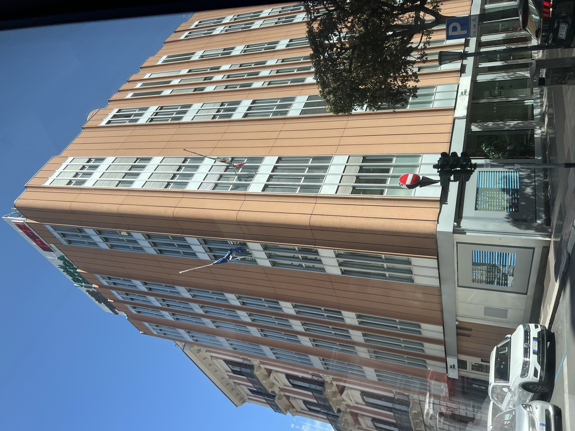 The image shows a multi-story building with a light orange facade and light gray window frames on a sunny day in Bari, Italy. The building has many windows arranged vertically in sections. At the top of the building, a red digital clock displays "15:49" and green lettering spells out "BANCA". Two flags, one blue and one red and white, are attached to the building. To the left of the orange building is a narrower, older building with a white and red facade, featuring balconies with dark railings. In the foreground, there are cars parked on a street, including a white Volkswagen SUV with license plate "FZ 085XI". A traffic light with red, yellow, and green signals is visible, as is a street sign with a blue "P" indicating parking and bicycle and pedestrian symbols. A dark green tree with dense foliage is on the right side of the image.