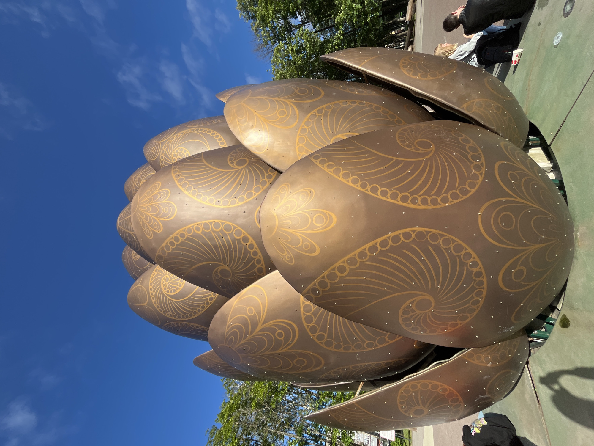 A large, metallic sculpture resembling an unopened lotus flower, with bronze-colored petals adorned with swirling gold patterns and small white studs, stands in a public area. The sculpture is viewed from a low angle, emphasizing its scale and detail. In the background, green trees with leafy branches and a bright blue sky with scattered white clouds are visible. To the right of the sculpture, two individuals, a woman with blonde hair and a man with dark hair, are seated on a green platform, facing away from the camera. The woman has a black backpack next to her and a red and white cup is visible on the ground. To the left, a portion of another person's black jacket and a shadow are visible, suggesting they are also present in the scene. A sign with text in a foreign language is partially visible in the far left background. The setting appears to be an outdoor park or plaza, with paved ground and some wooden elements suggesting seating or decorative features. This sculpture is located in Nizhny Novgorod, Russia.
