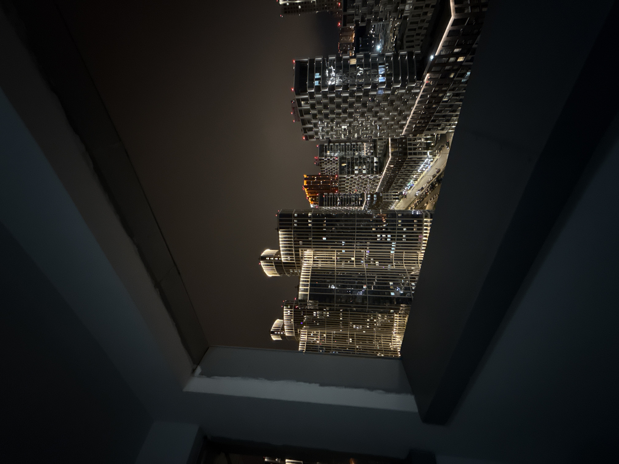The image displays a vibrant modern cityscape at night, viewed from an elevated position, likely from inside a building looking out through a window or opening. The scene is presented in an unusual vertical orientation, making the high-rise buildings appear to lie on their sides.
Numerous tall buildings dominate the frame, their facades brightly illuminated by a multitude of windows. These lights create a striking pattern against the dark night sky. Some buildings showcase distinct architectural features; for instance, a structure in the mid-ground glows with a prominent warm orange light, contrasting with the cooler white and yellow lights of surrounding towers. Other buildings have intricate, grid-like exteriors that catch the light.
At the base of some buildings, a street or road is visible, indicated by faint lines of vehicle lights, suggesting ongoing traffic. The sky above the city is dark, typical of nighttime, with a slight atmospheric glow from urban light pollution.
In the foreground, dark, angular architectural elements, possibly a window frame or parts of an interior wall and ceiling, frame the urban panorama. No specific human activity or individuals are discernible, and no visible text is present. The setting is an urban environment in Khimki, Russia, characterized by modern high-rise development.