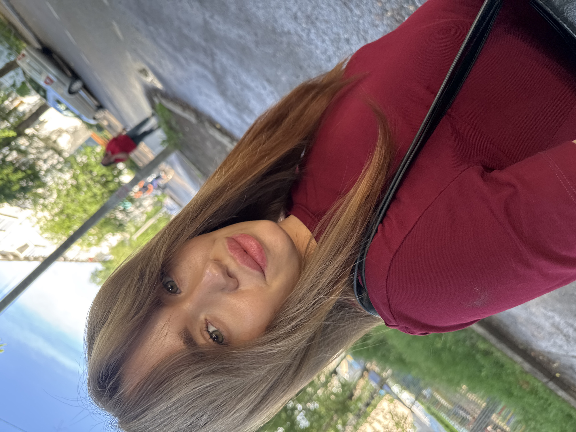 A young woman with long, light brown hair is captured in a selfie outdoors. She is wearing a dark red t-shirt and has a black strap visible across her chest, likely from a bag. The background suggests an urban environment with a paved street, a sidewalk, green foliage, and a building. A silver car is parked on the side of the street, and another person in a red top is visible in the distance, walking away from the camera. The sky is blue with some clouds. The image appears to be taken on a street in Chaikovsky, Russia.
