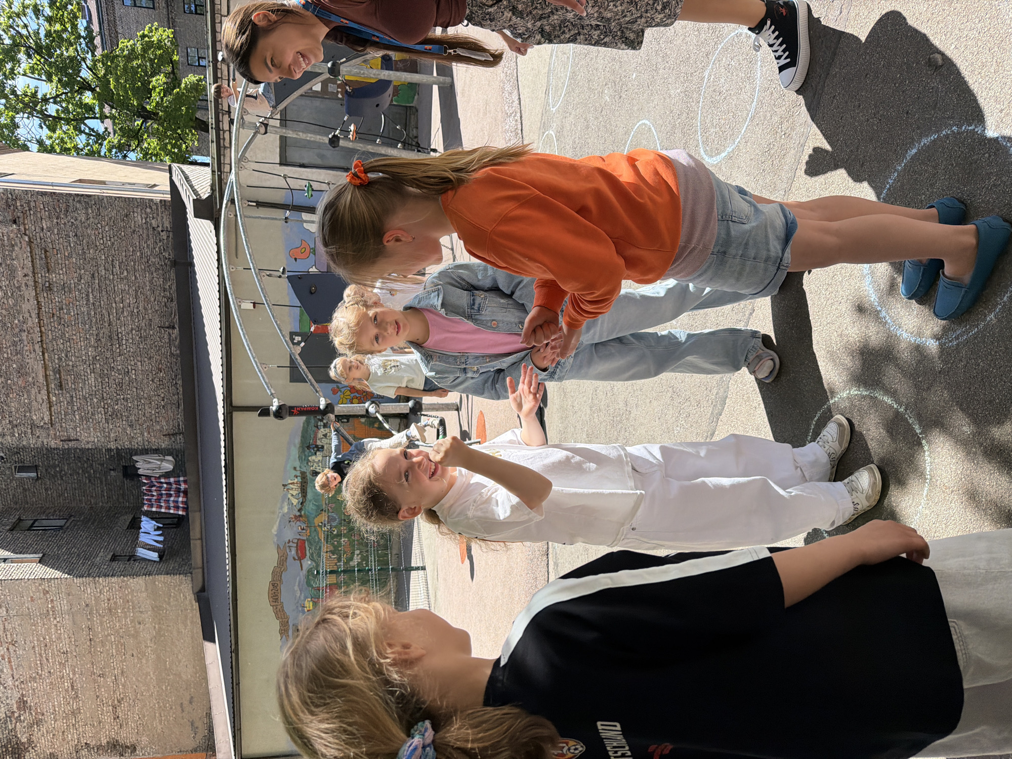 In a sunlit courtyard in Riga, Latvia, four children and a woman are engaged in an outdoor activity. The children, appearing to be of primary school age, are gathered in a circle marked with chalk on the asphalt. One girl in the foreground, facing away from the camera, wears a black and white t-shirt with "DEUTSCHLAND" visible on it and light-colored shorts. To her right, a girl in a white t-shirt and white pants has her arms outstretched and is laughing, with chalk dust on her hands and face. She is facing another girl wearing a pink t-shirt and a denim jacket, who is also looking towards the first girl. A fourth girl in an orange sweatshirt and denim shorts stands facing the other two, with her hands clasped in front of her. Behind them, a woman with dark hair tied back smiles broadly, wearing a brown top and a patterned skirt. In the background, a multi-story brick building with laundry hanging from a clothesline is visible, along with playground equipment and a mural on a wall. The scene suggests a playful and interactive learning environment.