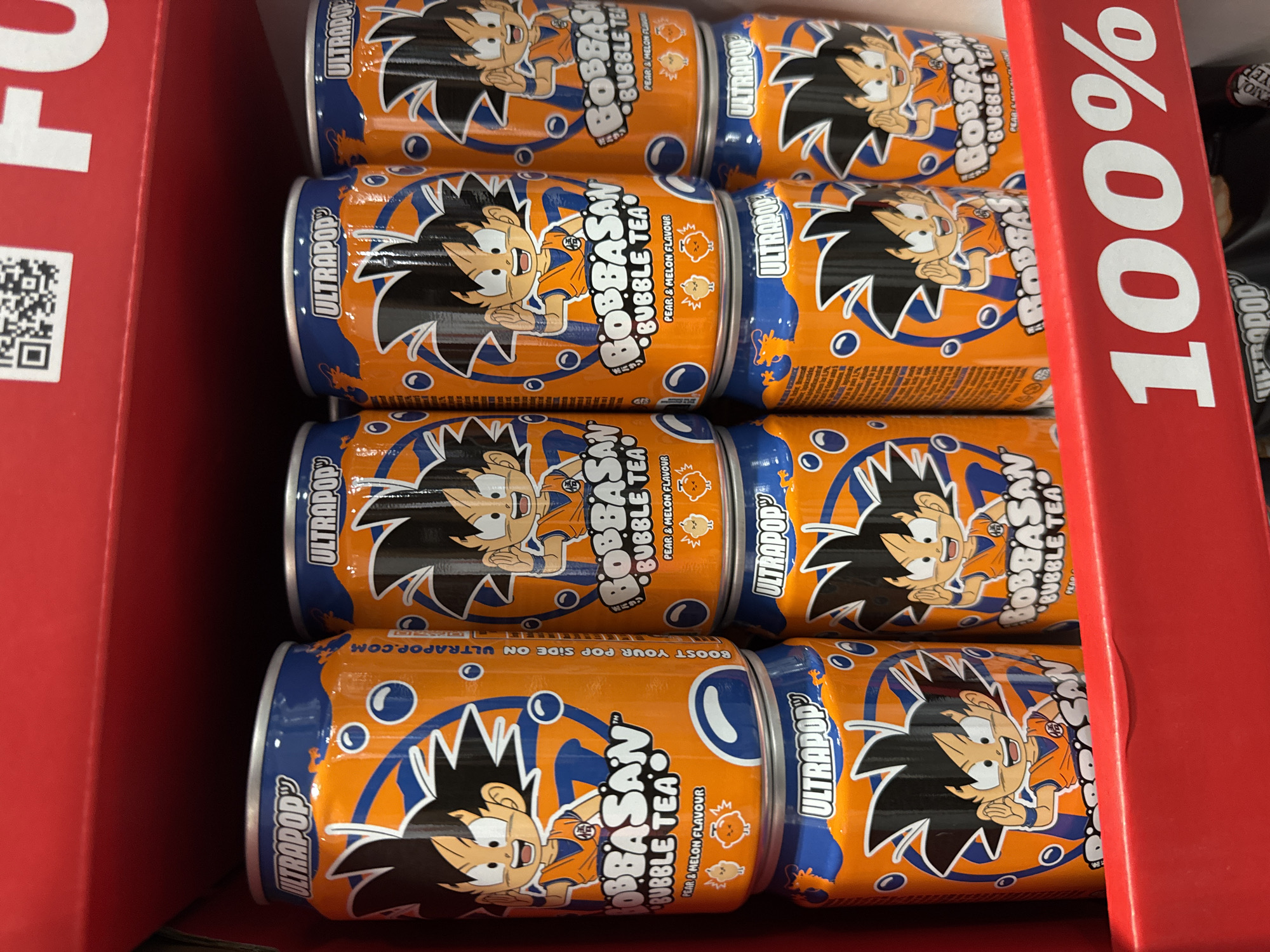 The image shows a shelf in a retail environment, likely a convenience store or supermarket, displaying cans of "BOBBASAN BUBBLE TEA" in various flavors. The cans feature an animated character, resembling Goku from Dragon Ball Z, in an orange gi. The text on the cans includes "ULTRAPOP" and "BUBBLE TEA PEAR & MELON FLAVOUR." A prominent red strip with "100%" printed in large white font is visible at the bottom of the image. The background is out of focus but suggests a retail display with a red backdrop. The overall impression is one of a product promotion or display in a store. There is no indication of the specific location being in Sofia, Bulgaria.