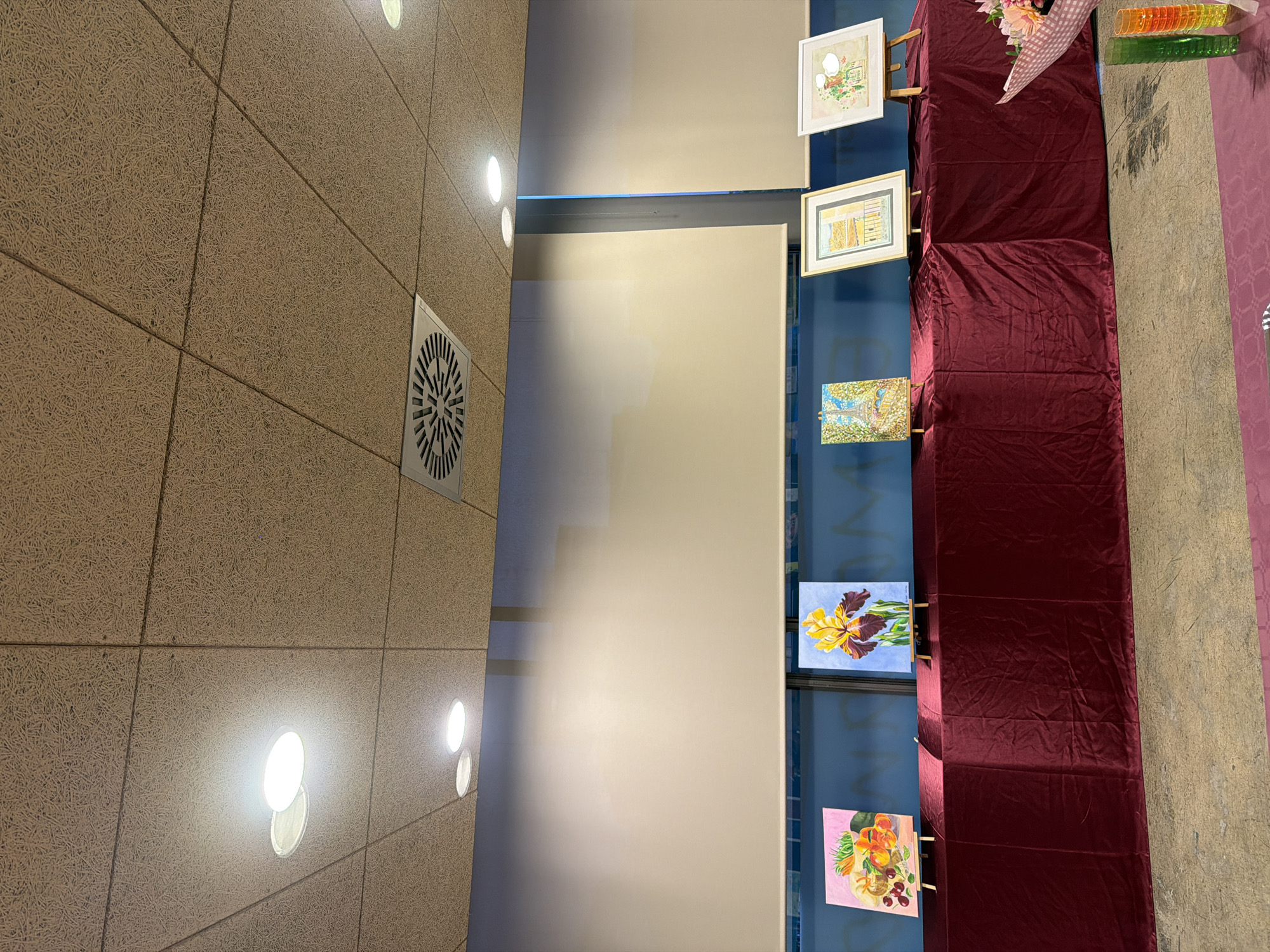 The image shows an indoor exhibition with several paintings displayed on easels atop a burgundy velvet-covered table. From left to right, the paintings depict a still life of fruits, a single iris flower, a view of the Eiffel Tower, a balcony scene, and a floral arrangement. The room has a textured ceiling with recessed lighting and air vents. A large, light-colored roller blind covers the upper portion of the wall behind the table. The floor appears to be a concrete or tiled surface. There are no people visible in the frame. The scene is likely from a local exhibition or event in Elche/elx, Spain, possibly a community center, gallery, or school hall, given the simple presentation.