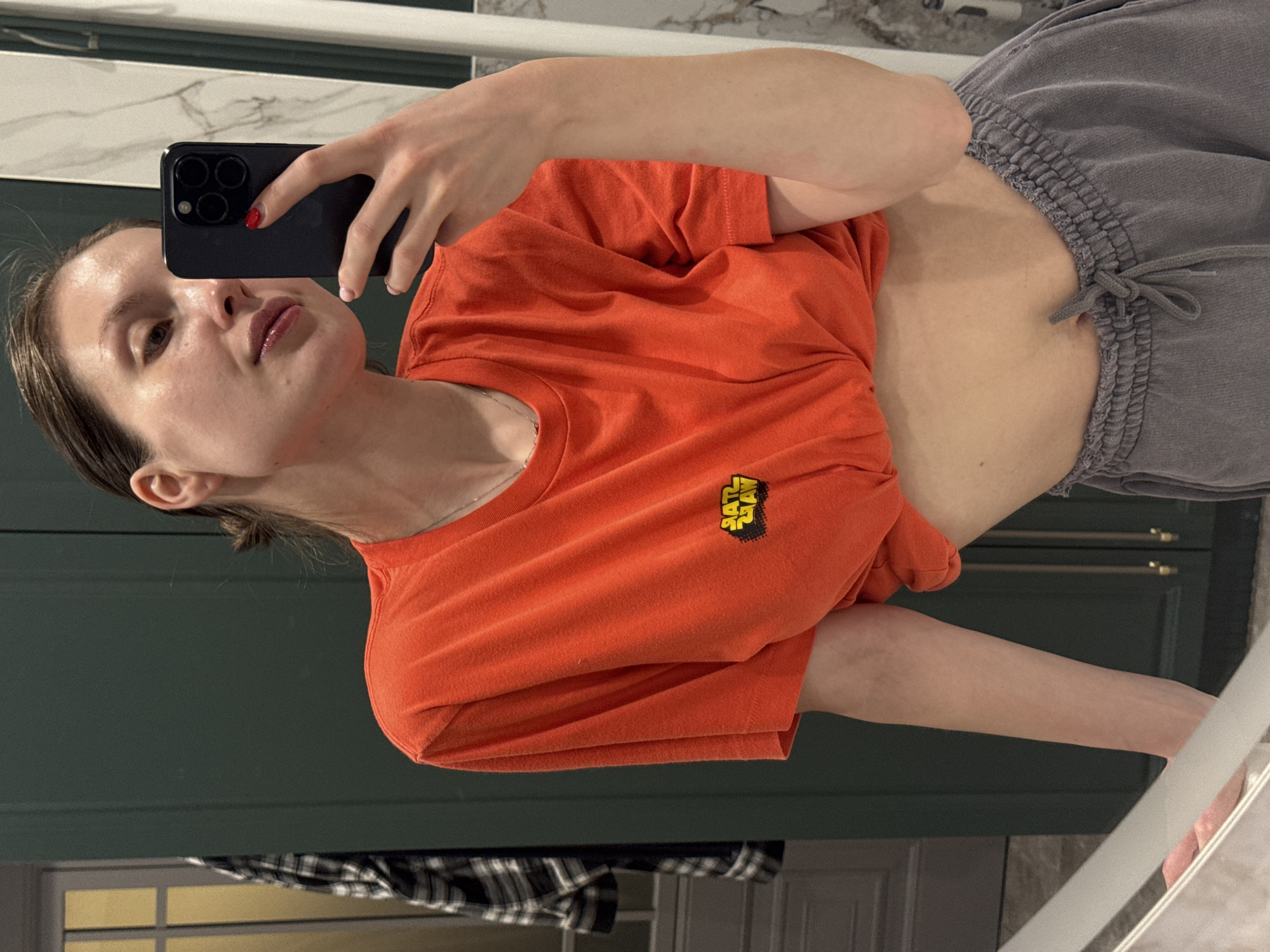 A woman is taking a selfie in what appears to be a bathroom. She is wearing an orange t-shirt with a Star Wars logo and grey sweatpants. She is holding a black smartphone with a red nail visible on her index finger. The background shows a marble countertop, a white-striped wall, and a dark green cabinet. The location may be Tomilino, Russia.