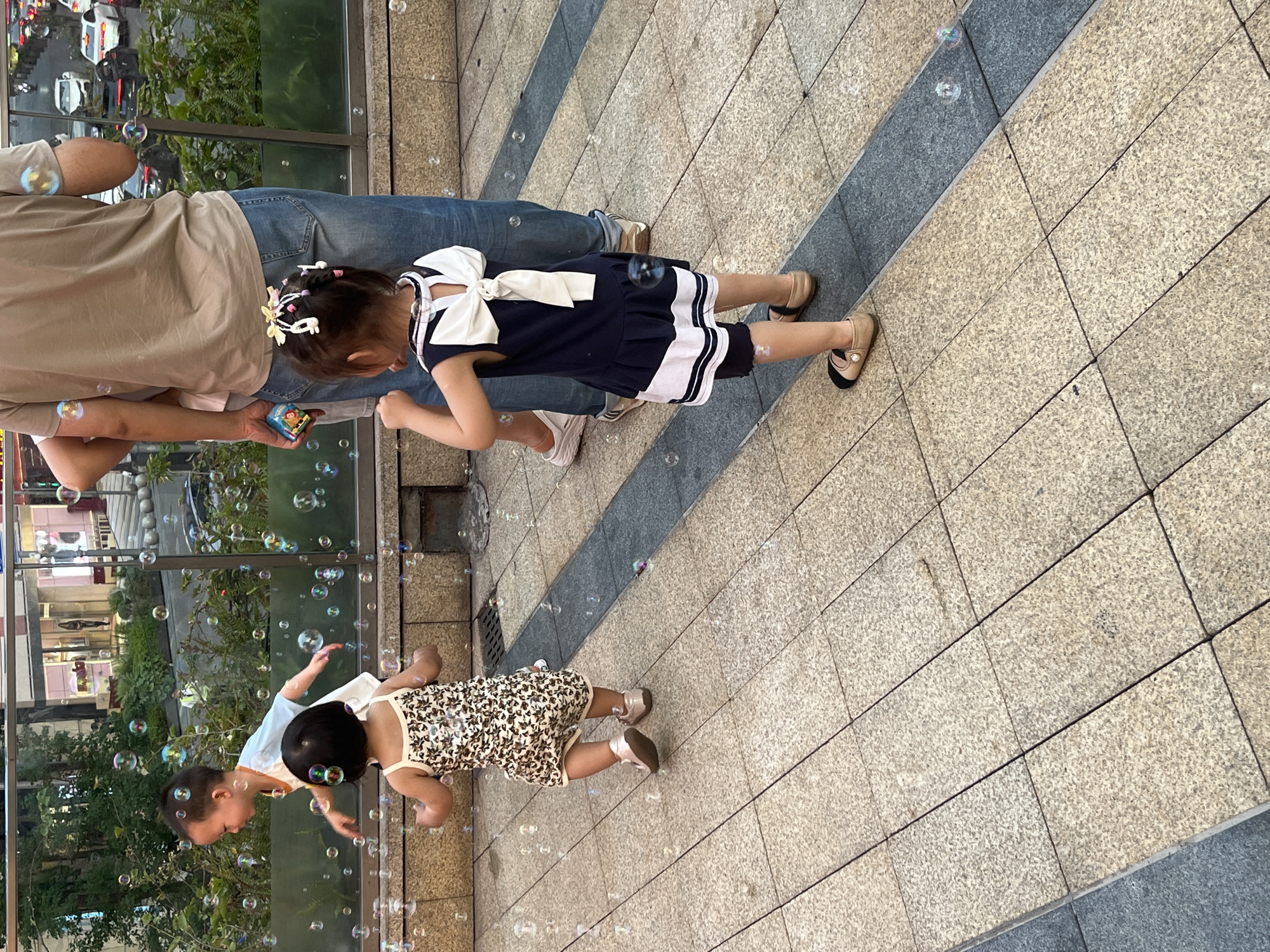 The image depicts an outdoor urban scene in Yuzhong District, China, featuring four individuals on a paved pedestrian area interacting with numerous iridescent bubbles.
On the left, a young boy with short dark hair, wearing a white t-shirt with an orange collar, is captured in profile, facing right, with his arms slightly raised, appearing to move towards the bubbles. In front of him, a toddler, seen from the back, wears a light-colored sleeveless dress with a dark, possibly floral, pattern and shiny light-colored flat shoes. This child is in motion, walking or running towards the right side of the frame.
On the right side of the frame, a young girl, also seen from the back, stands facing away from the viewer. She has dark hair styled in pigtails adorned with small yellow flower clips and white, possibly plastic, clips. She wears a navy blue dress with white horizontal stripes on the skirt and a prominent white bow on the back over a cutout. She is wearing light-colored flat shoes with black toes. Behind and to the left of this girl, the lower back and legs of an adult are visible, wearing a light brown t-shirt and blue jeans. This adult holds a small, colorful rectangular object, likely a bubble solution container or toy, in their left hand. Partially obscured next to this adult is another individual's arm and hip.
The ground is paved with light brown, square granite-like tiles, which transition to a darker grey strip in some areas. Numerous bubbles of varying sizes are suspended in the air across the entire scene. In the background, behind a transparent glass barrier, there is green foliage from bushes and trees, beyond which a multi-lane street with vehicular traffic (cars and a motorcycle) and modern buildings are visible under daylight conditions. A small metal drainage grate is present on the paving in the mid-ground.