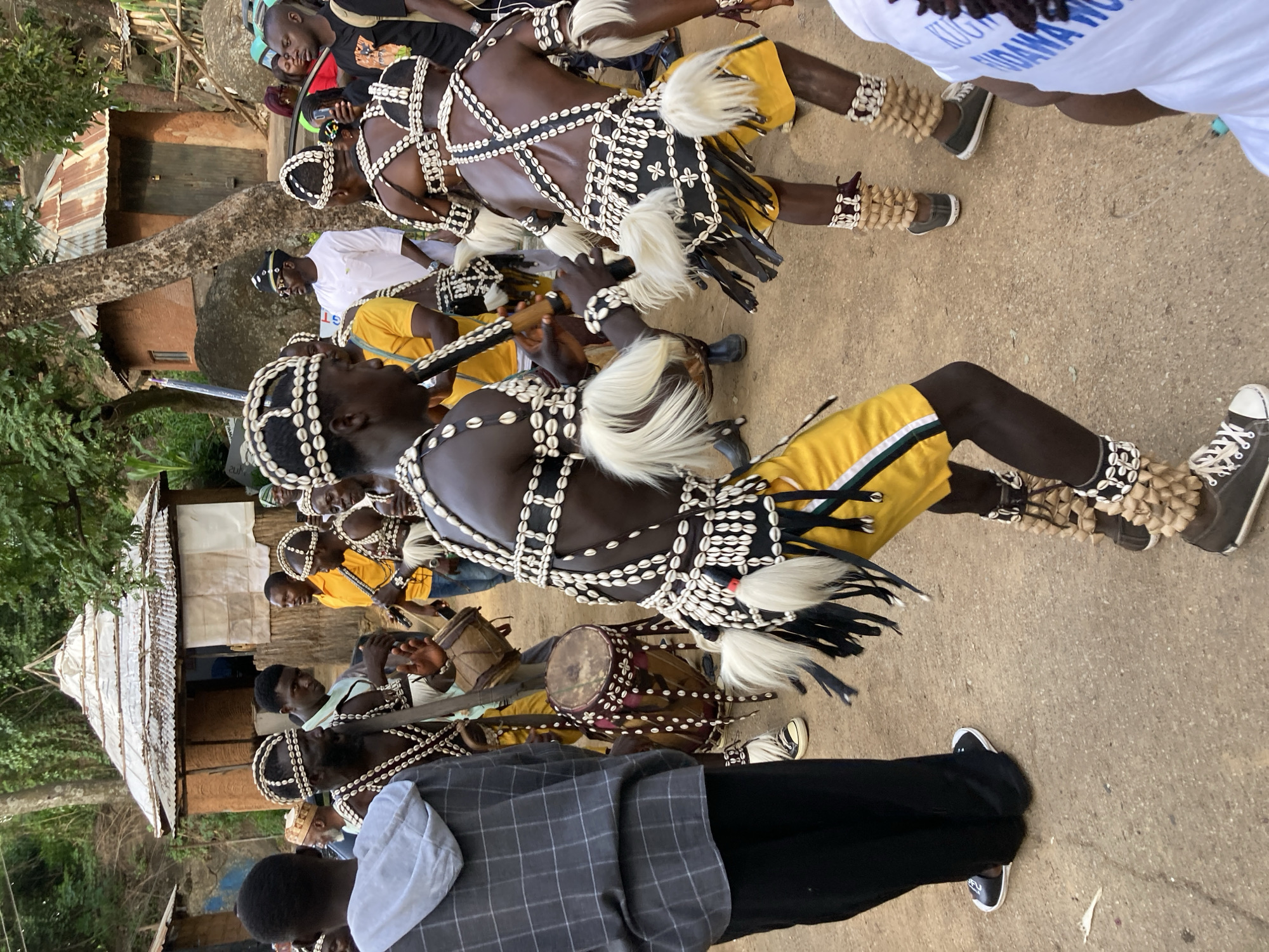 In an outdoor scene at a village setting in Jos, Nigeria, several individuals are engaged in a cultural performance. The foreground is dominated by several men adorned in traditional attire. These performers wear beaded headwear, necklaces, and armbands made of white shells and dark beads. Their outfits include dark loincloths decorated with shell patterns and prominent white fur tufts. Some also wear yellow shorts or wraps. Two individuals in the foreground are playing drums, one a larger cylindrical drum and another a smaller hand drum. Another performer is playing a flute-like instrument. In the background, a traditional hut with a conical roof made of corrugated metal sheets and thatched walls is visible. Other structures, including a more rectangular building with a wooden door, are also present. A diverse group of onlookers, some in casual Western clothing and others in traditional attire, observe the performance. The ground is a dirt path. The overall atmosphere suggests a cultural event or festival.