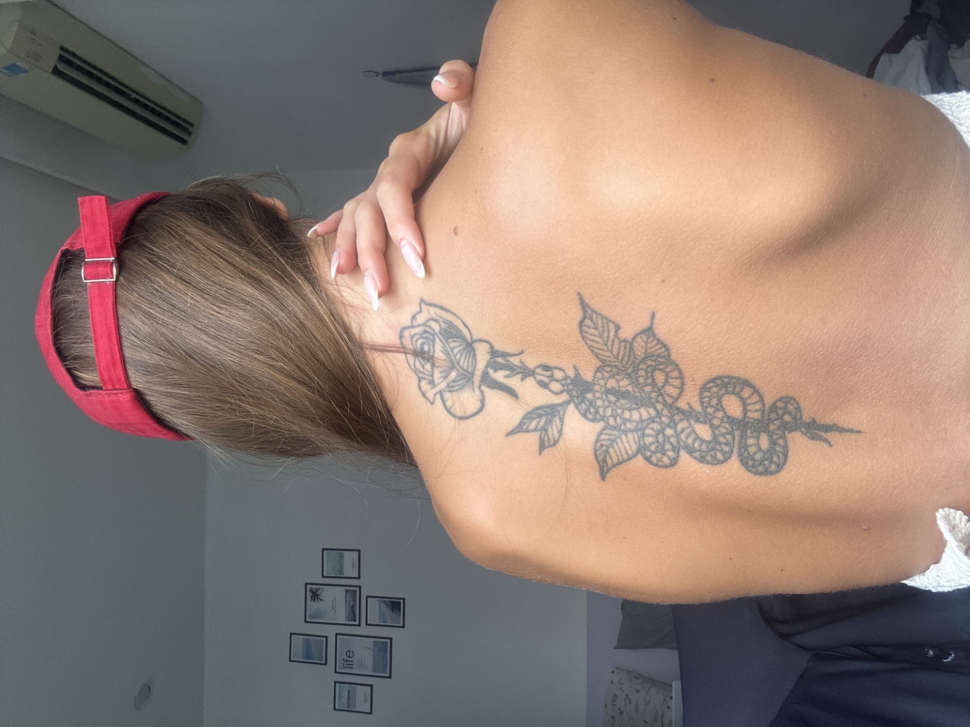 A woman is seen from the back, showcasing a tattoo of a rose and snake design on her upper back. She is wearing a red baseball cap. Her hand is raised towards her neck. The background shows part of a room with a window, a wall with framed pictures, and an air conditioning unit mounted near the ceiling. There is no information in the photo that indicates its location in Đà Nẵng, Vietnam.
