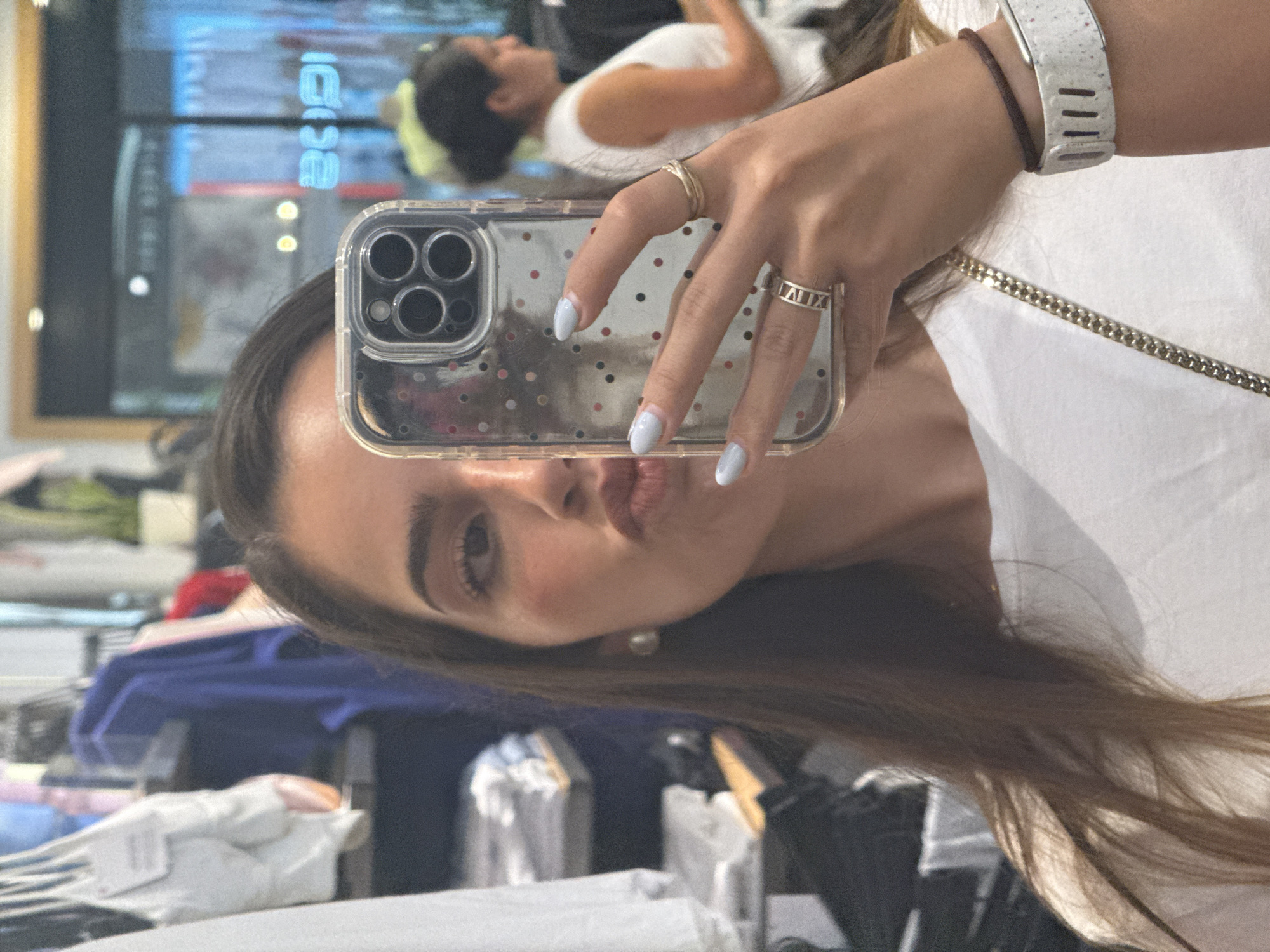 A selfie-style shot, likely taken in a mirror, features a woman holding a cell phone with a clear case decorated with small red, black, and gray dots. The phone is positioned to obscure the central part of her face. Her hair is brown, and she is wearing a white shirt with a gold chain visible at the neckline. Her fingernails are painted a light blue color. She is wearing two rings on her right hand; one is a simple band, and the other features Roman numerals. A fitness tracker is on her wrist, partially obscured.
In the reflection of the mirror behind her, another woman with her hair in a bun and wearing a white shirt is visible. The background shows clothing racks with various garments. Faint reflections of blue lights are also visible.
The location appears to be a clothing store in Puebla, Mexico.