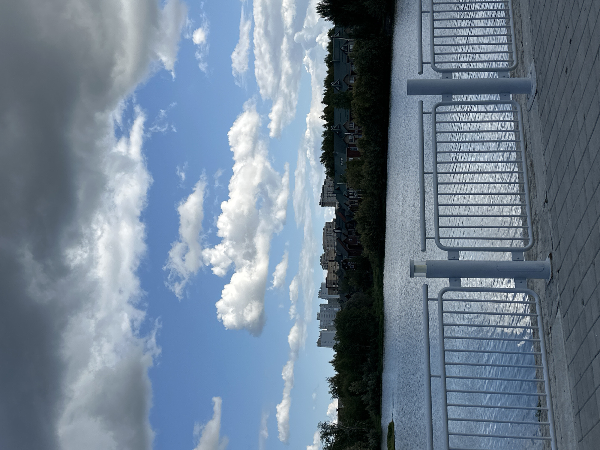 The image depicts a waterfront scene in Surgut, Russia, likely a park or embankment area. In the foreground, a paved walkway with a grey, rectangular paver pattern is visible, leading to a white metal railing. This railing, composed of vertical bars and horizontal top rails, runs along the edge of the walkway, separating it from a body of water. The water, appearing calm with gentle ripples, reflects the light from the sky.
Beyond the water, a dense line of green trees forms the middle ground. Behind the trees, a cityscape emerges. On the left side of the image, several tall, multi-story residential buildings with light-colored facades are prominent. To the right of these buildings, a cluster of houses with dark green roofs and traditional architectural elements can be seen, interspersed with more trees.
The sky is a dominant feature of the image, with a dramatic mix of blue sky and large, fluffy white and dark grey clouds. The overall lighting suggests it is daytime, with some areas illuminated by sunlight and others shaded by the clouds. The scene conveys a sense of a developed urban area adjacent to a natural water body, with the sky contributing to a dynamic and somewhat overcast atmosphere. There are no people or animals visible in the image.