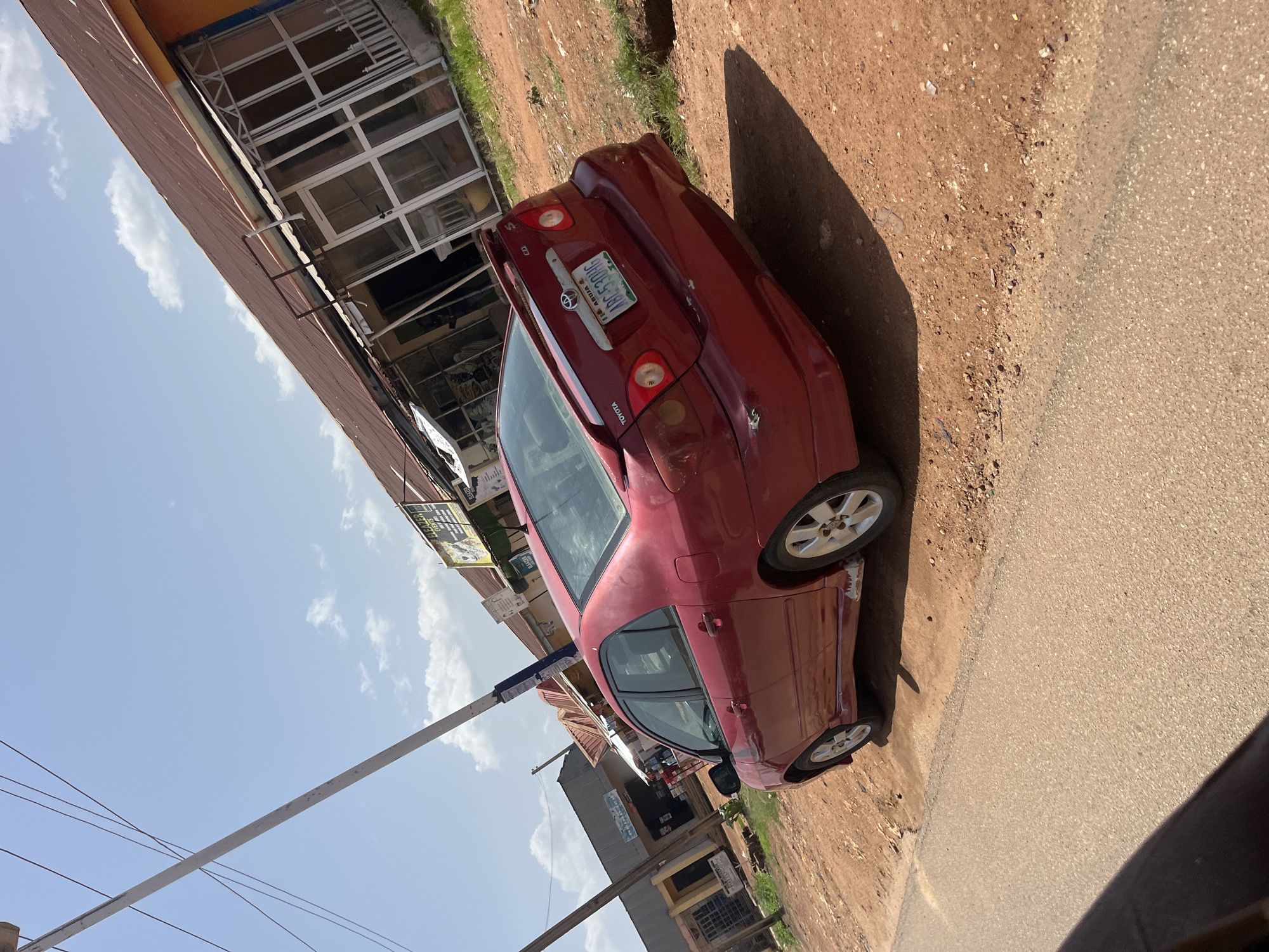 A red Toyota sedan is parked on a dirt shoulder next to a paved road. The car is seen from a low angle, tilted on its side. The license plate reads "ABE 530HG". In the background, there is a building with windows and a brown metal roof, possibly a shop. There are also some signs visible on the building, one of which appears to say "DEALER". The sky is blue with some white clouds. The sun is casting a shadow from the car onto the ground. The overall impression is of a street scene in a town, likely during the day. The scene is outdoors in what appears to be a warm climate, given the lighting and clear sky. There are no people visible in the image. The setting appears to be a commercial area with businesses lining the street.