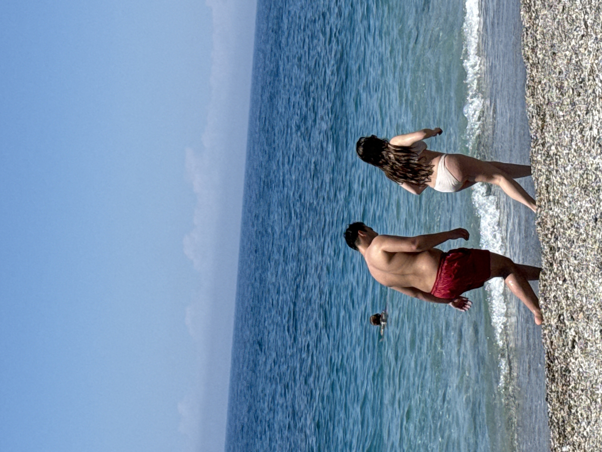Here is a detailed description of the image:
**Location:** The image depicts a beach scene.
**People:** There are three people visible:
1. A woman with long brown hair, wearing a white bikini, is standing at the edge of the water.
2. A man with a bare torso and red shorts is standing beside her, also at the edge of the water.
3. Another person, possibly a child, is swimming in the water.
**Environment:**
* The beach has a shoreline of small stones or pebbles.
* The water is clear blue, with white waves breaking near the shore.
* The sky is bright blue, with a slight haze near the horizon.
**Actions and Interactions:**
* The man and woman appear to be wading in the water, standing at the edge of the shore.
* The swimmer is active in the water, further from the shore.
**Objects:**
* There are no other objects of note in the image.