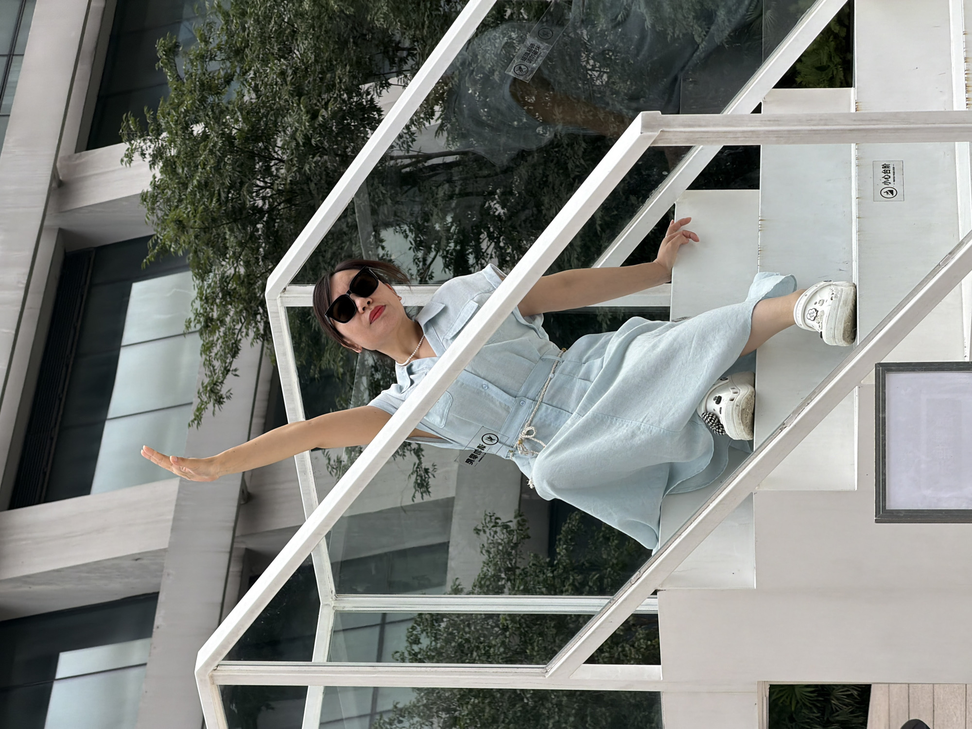 A woman in a light blue dress and sunglasses is sitting on a white outdoor staircase, raising her right arm. She is wearing white clogs with black and white patterns. The staircase has white railings and glass panels. Behind the woman, a modern building with large windows and trees can be seen. The scene appears to be in Yuzhong District, China, possibly near a contemporary architectural structure.