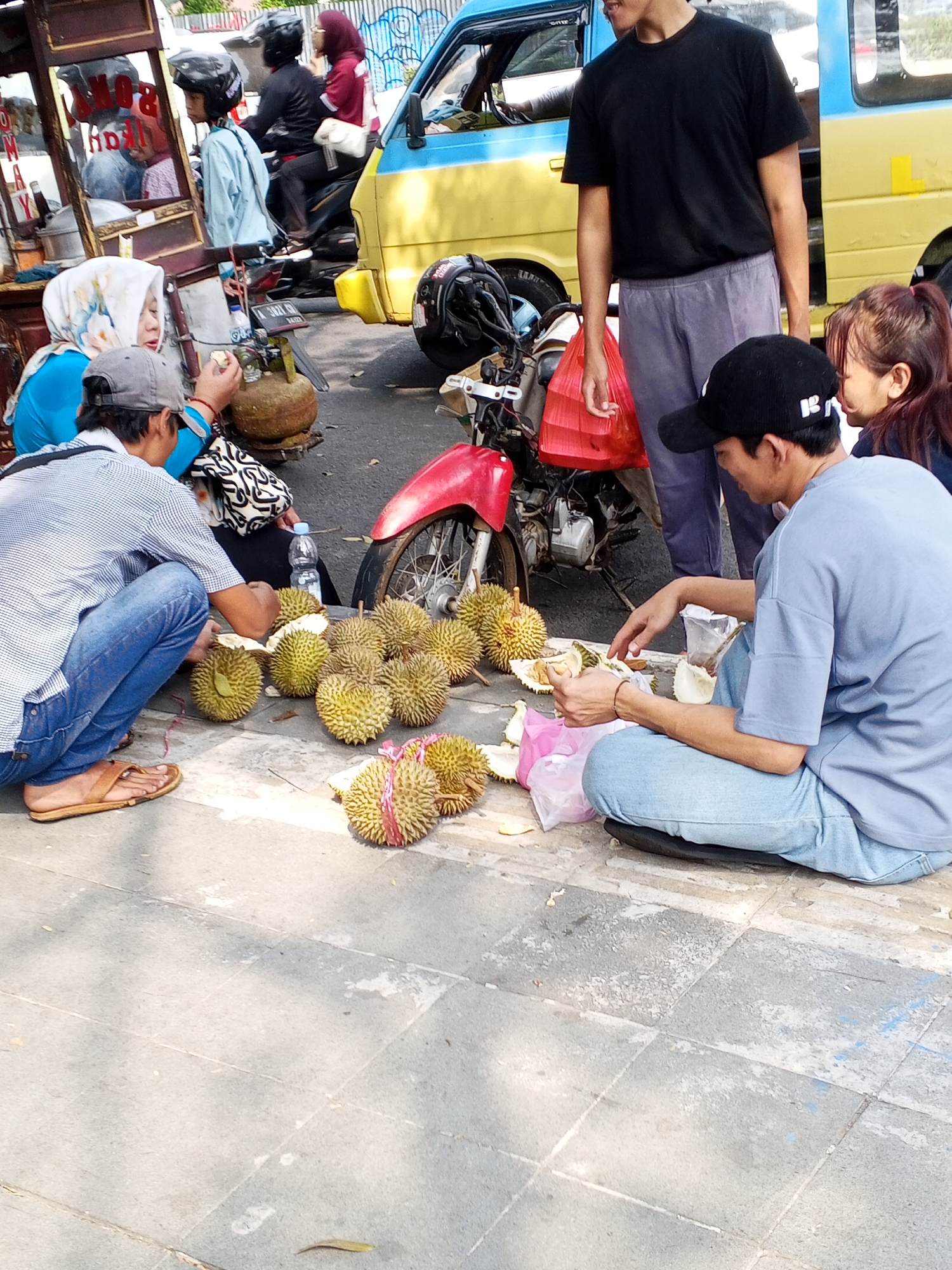 The image depicts a street scene in Serang, Indonesia, with a group of people interacting near a street vendor's cart and a motorcycle. The foreground shows a man wearing a blue t-shirt and jeans, sitting on the ground and eating durian. He is surrounded by several durian fruits, some of which have been opened. To his left, a man in a striped shirt and jeans is also seated, preparing to eat durian. A woman wearing a blue hijab and blue top is to his right, also near the durian. Behind them, a yellow and blue vehicle, possibly a small truck or van, is parked. A red motorcycle with a red fender is also present. Other individuals are visible in the background, including a person wearing a black t-shirt and gray pants, who appears to be a vendor, and several others on motorcycles. The ground is paved with gray tiles, and some debris is scattered around. The overall atmosphere suggests a casual street food setting.