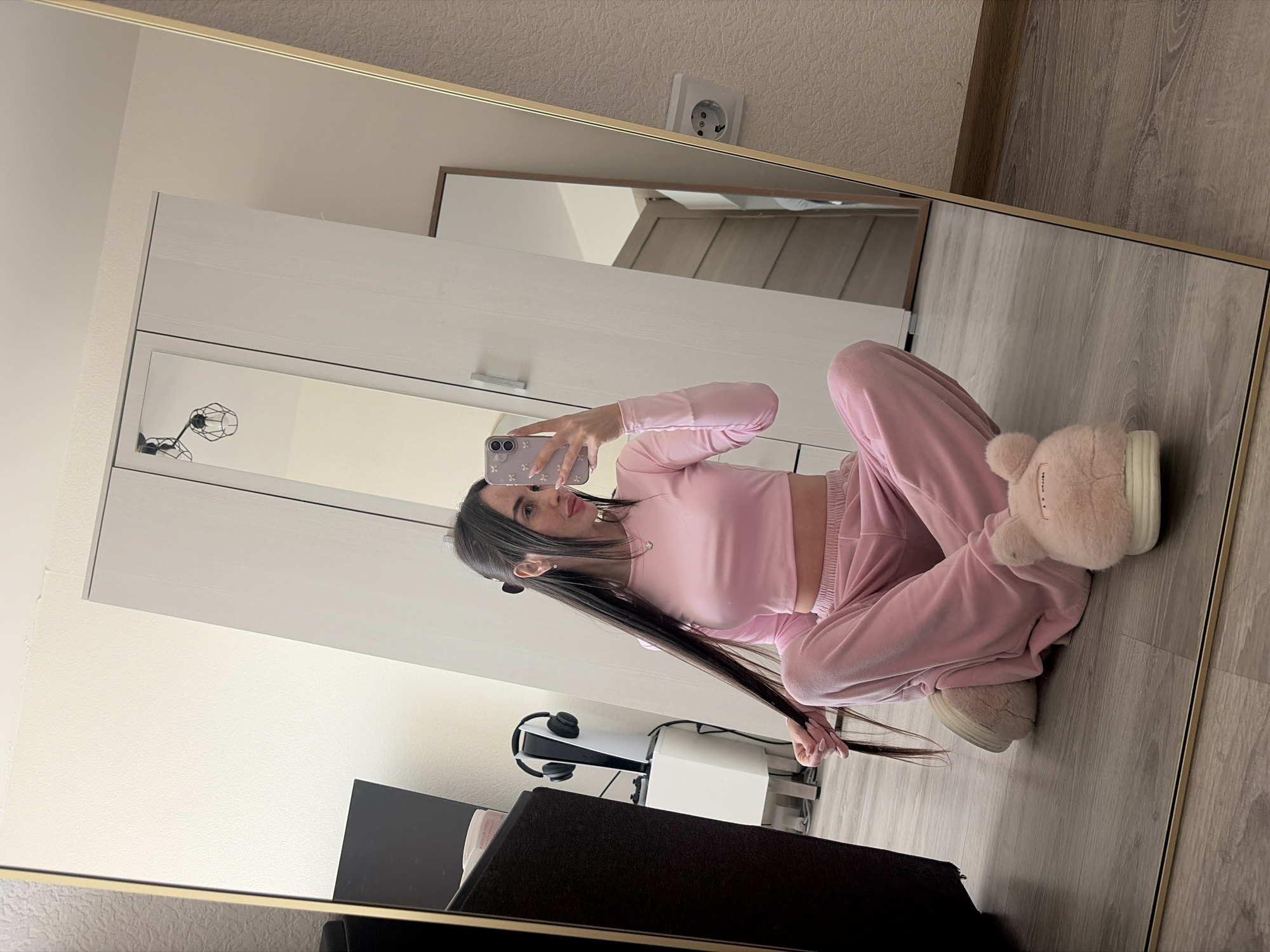 A young woman is taking a selfie in a mirror. She is wearing a pink long-sleeved top and pink pants, along with pink slippers. She is sitting on a gray wood floor. The background includes a white wardrobe, a smaller mirror reflecting a slanted ceiling, an electrical outlet, a black television, a white table with a PlayStation 5 and headphones, and a wall. The location appears to be a room within a domestic residence. There is no indication that this image was taken in Saint Petersburg, Russia.