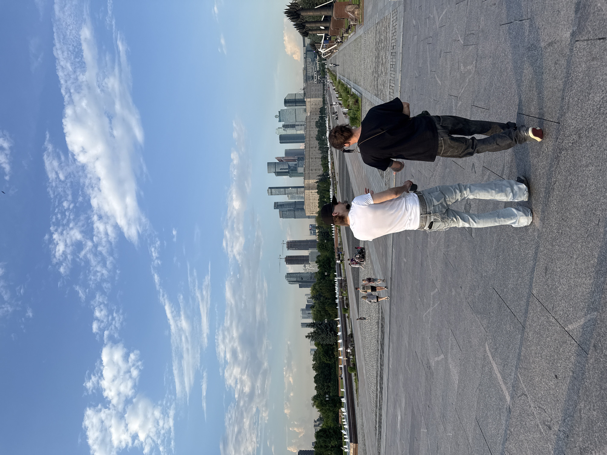 In Moscow, Russia, two individuals walk away from the camera on a wide, paved plaza. The person on the left wears a white t-shirt and light-wash jeans with a chain belt. The person on the right wears a black t-shirt and dark jeans. Behind them, a row of fountains runs parallel to a tree-lined area and a long, low building. In the distance, a cluster of modern skyscrapers dominates the skyline under a partly cloudy blue sky. The plaza is made of large, grey stone slabs with visible lines and some scuff marks. Other people are scattered throughout the plaza, some walking, some with a stroller, and a few are seated on benches. A single bird is visible in the sky.