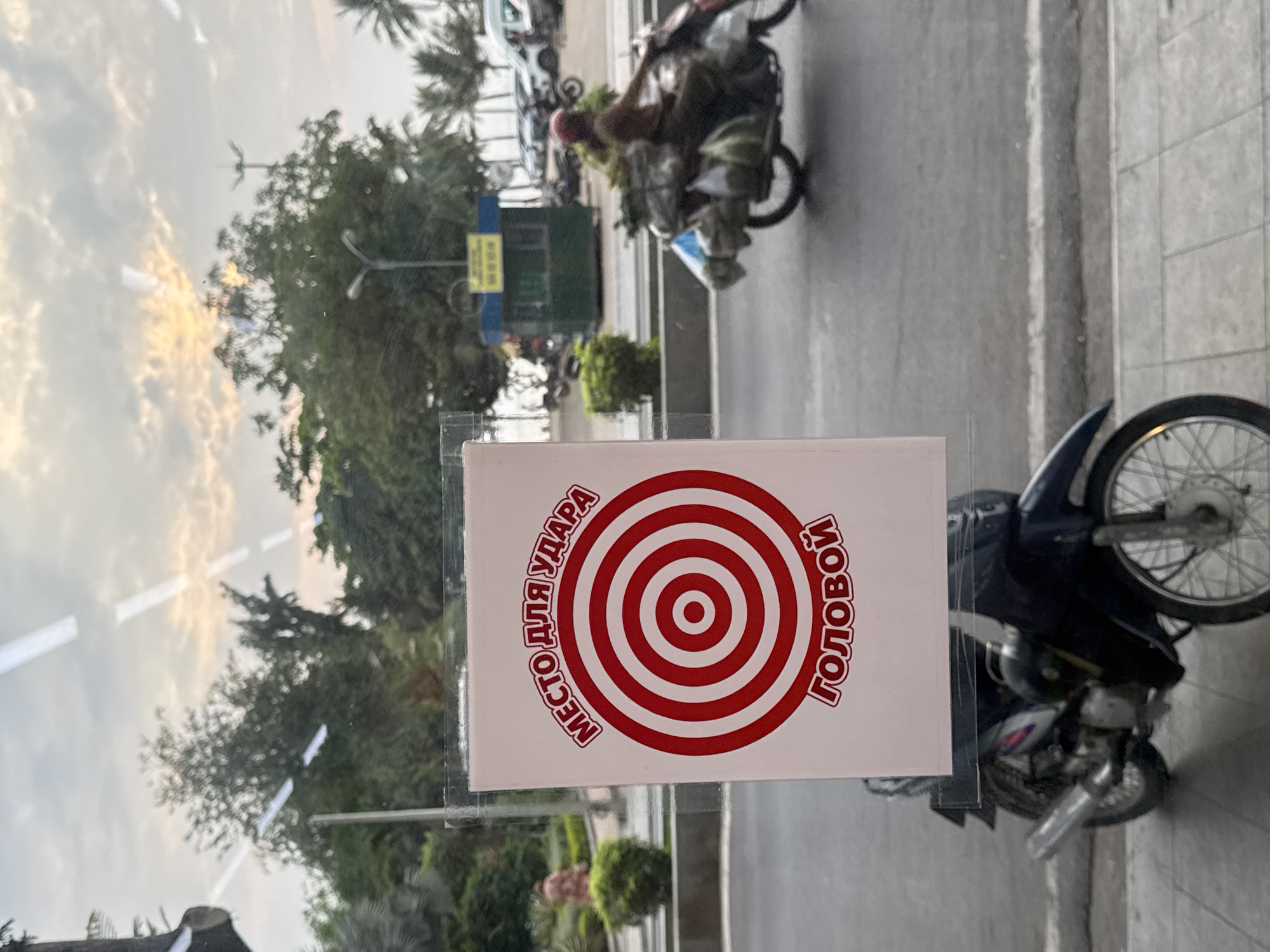 The image shows a view through a glass window with a sign attached to it. The sign is white with a red bullseye design and Cyrillic text around it. The text is oriented vertically, running along the left and right sides of the bullseye.
Through the window, a street scene is visible. On the right of the window, a parked motorcycle is partially visible. Beyond the motorcycle, the street stretches into the distance. A person on a heavily laden motorcycle is traveling along the street. Further back, a green kiosk or stall can be seen. The background shows trees and a sky with cloud formations, suggesting the time of day may be near sunset or sunrise. The scene seems to be taken in a city ward in Nha Trang, Vietnam.